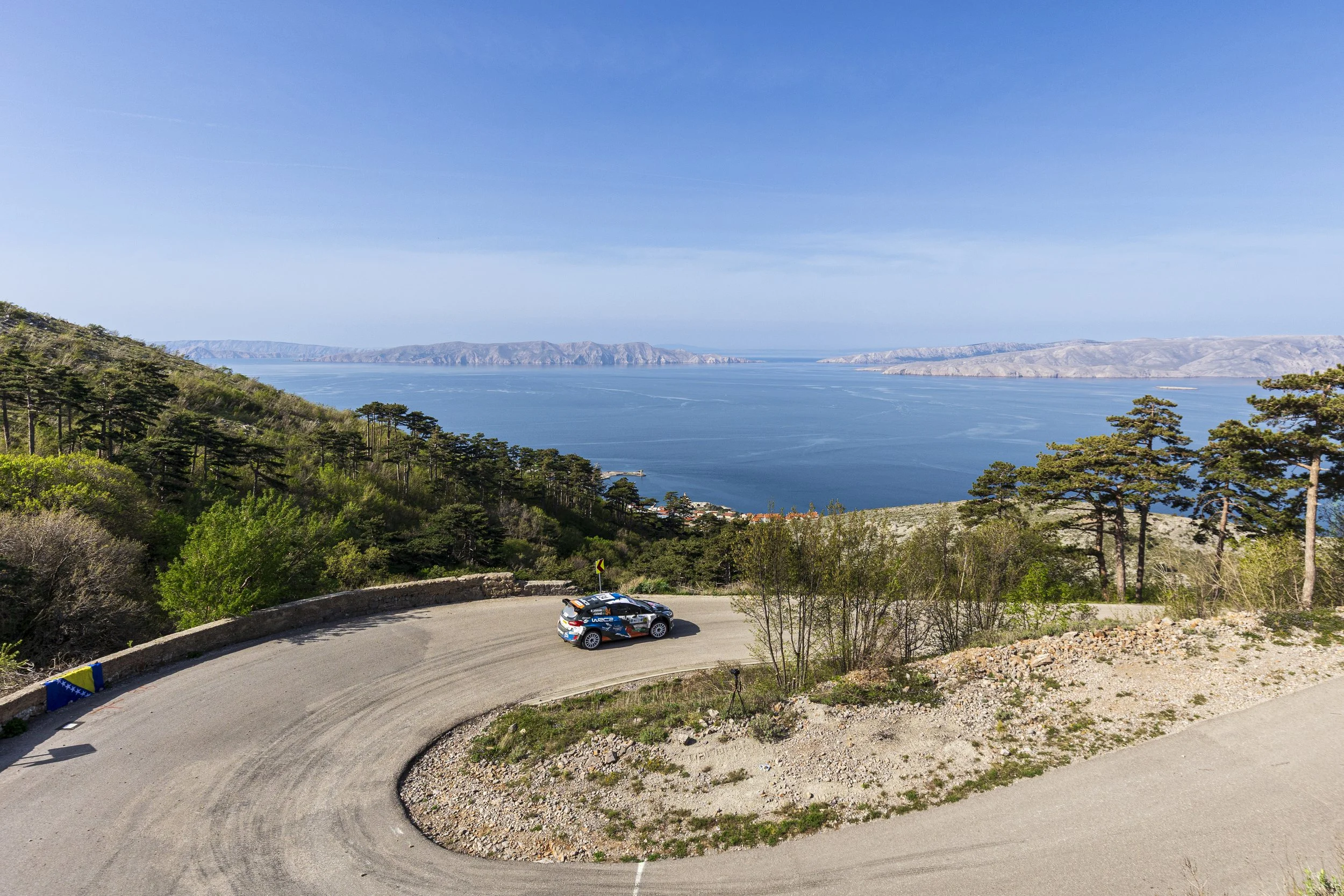 A rally car driving on a curvy mountain road overlooking a large lake or sea with mountains in the background and trees in the foreground.