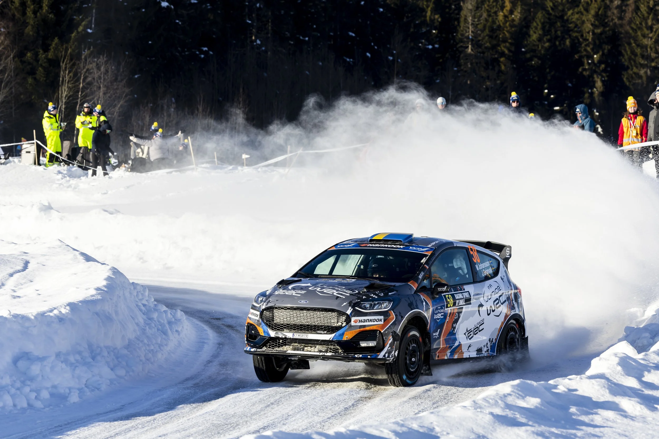 A race car on a snowy track during a winter rally race, with spectators watching from the side and snow-covered trees in the background.