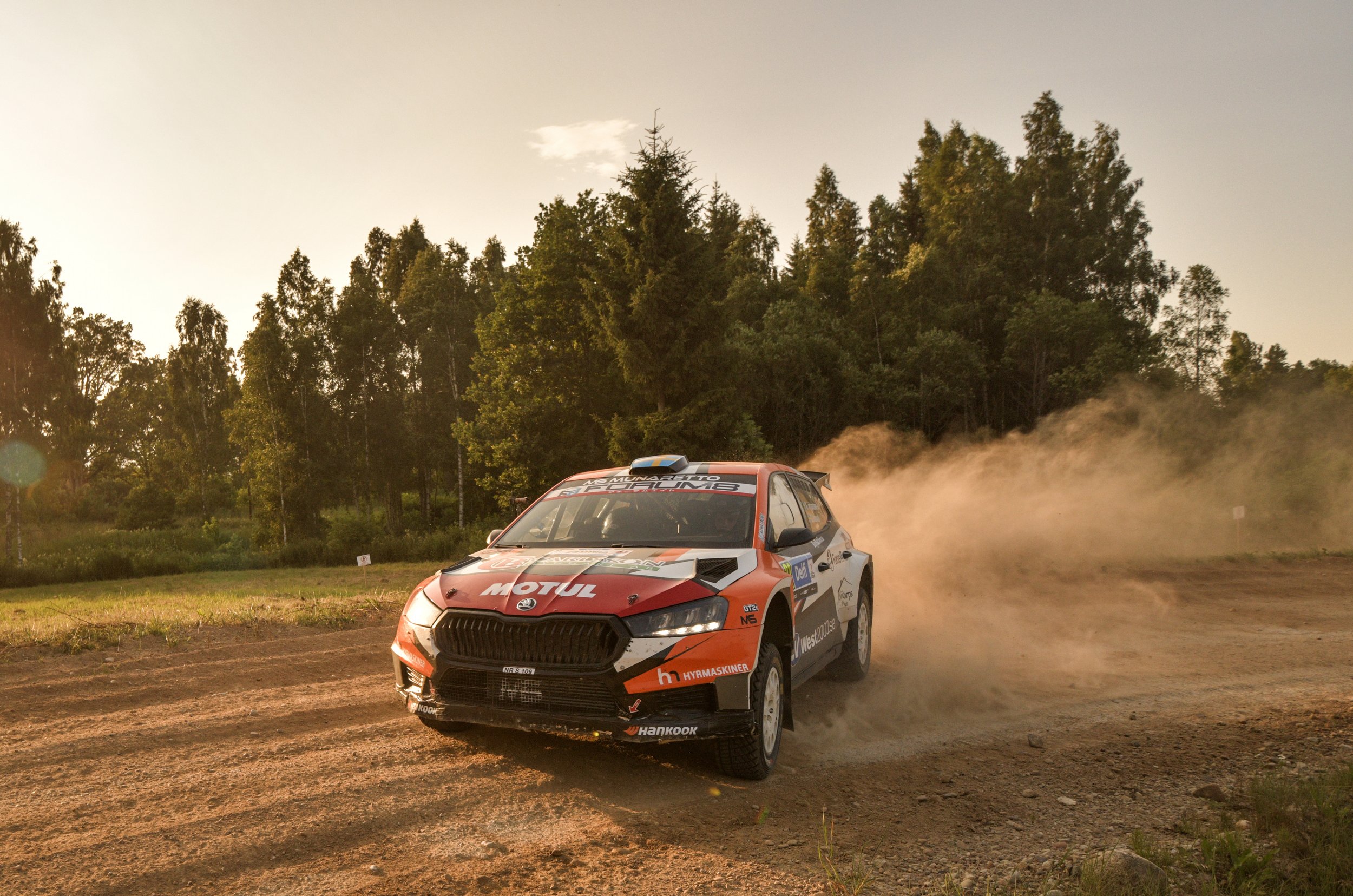 A rally car racing on a dirt track with dust clouds behind it, set against a backdrop of green trees and a partly cloudy sky during sunset.