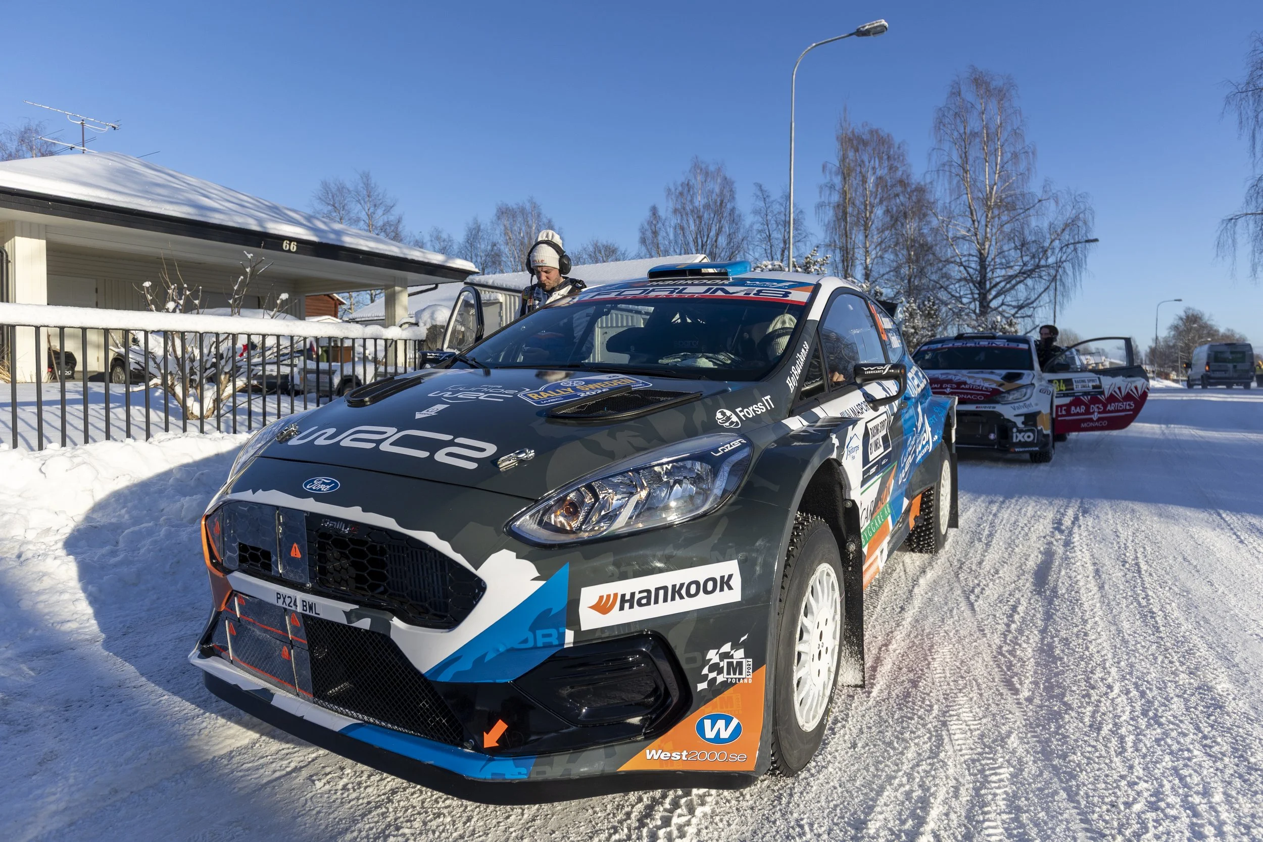 A rally car parked on snowy ground with a person wearing headphones and a beanie standing beside it. The car features sponsor logos and a racing number, and is in front of a residential house with trees and a street lamp in the background.
