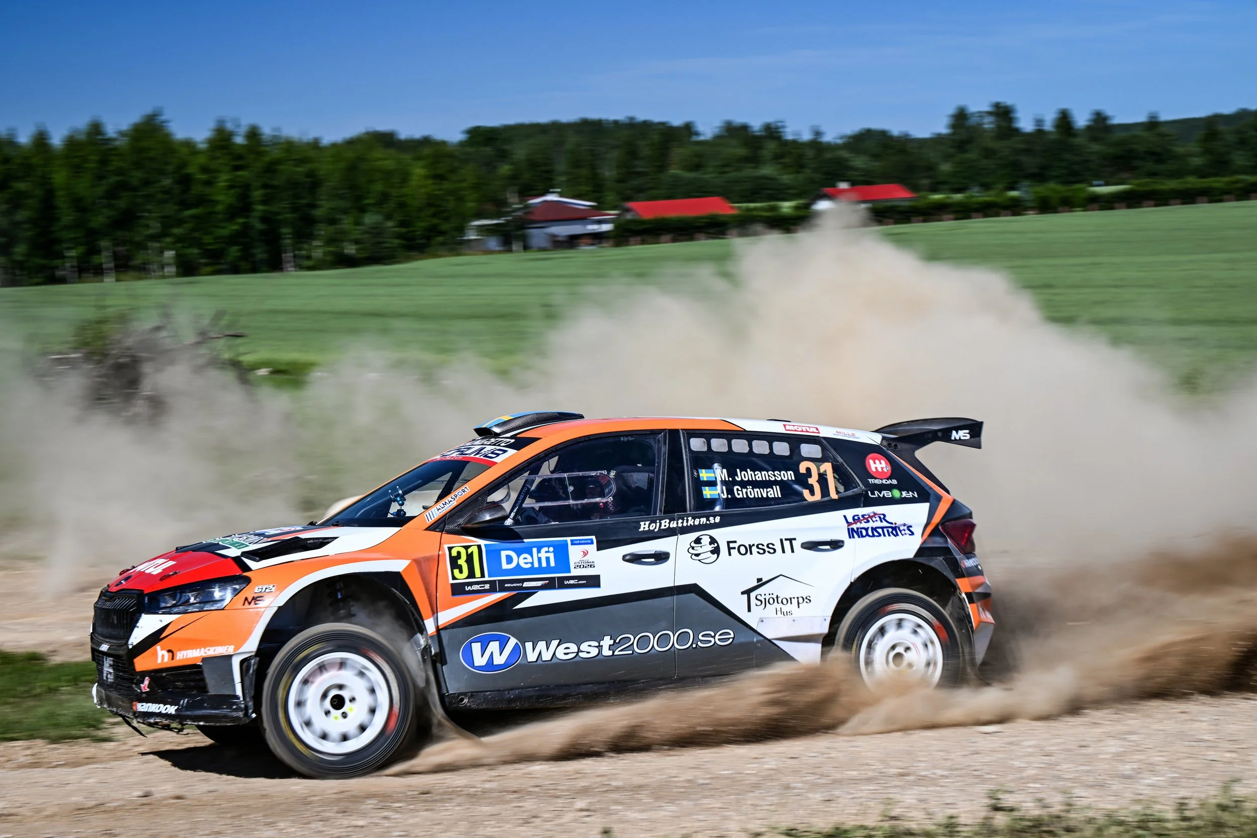 A rally car driving on a dirt road kicking up dust, with a green landscape and trees in the background.