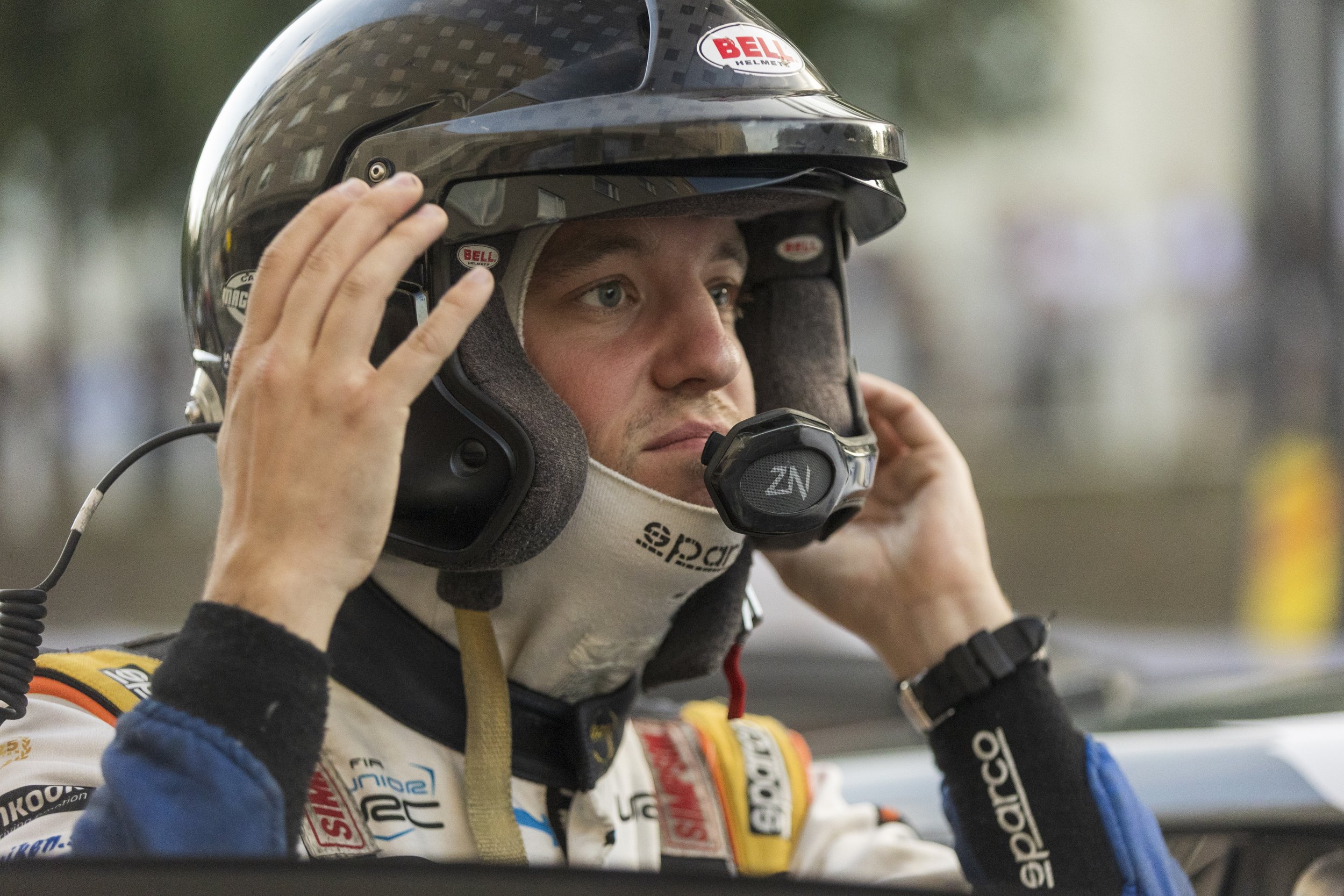 A race car driver wearing a black Bell helmet with a carbon fiber pattern, adjusting his helmet strap, dressed in a racing suit with various sponsor logos, in a racing pit or garage setting.