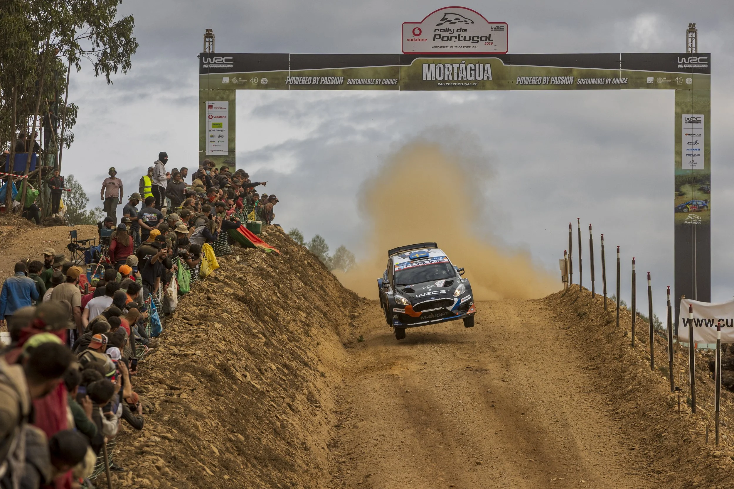 A rally car airborne on a dirt track during a race, with a large crowd of spectators on the left side and banners overhead, in Mortágua, Portugal.