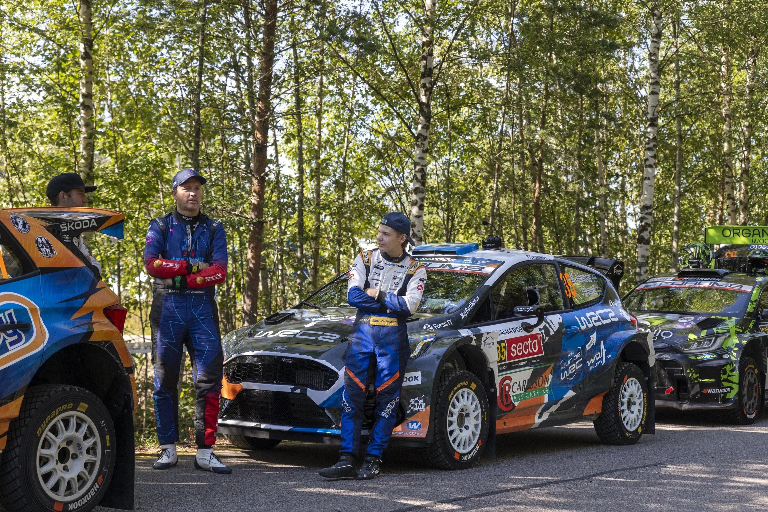Race car drivers standing beside their rally cars in a wooded area, preparing for a race. The cars are decorated with sponsor logos and numbers, and the drivers are dressed in racing suits.
