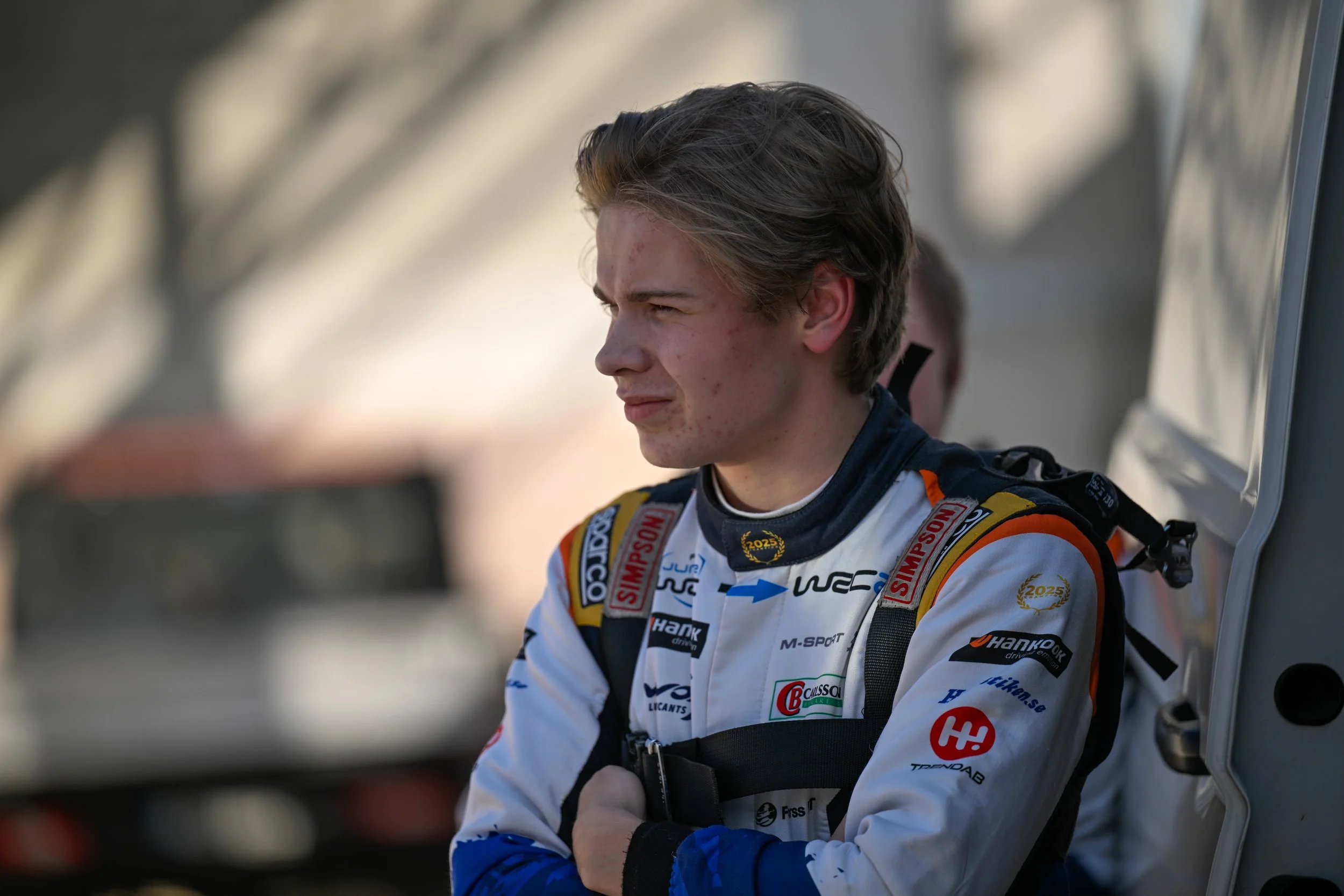 Race car driver in a white racing suit with various sponsor logos, standing next to their vehicle, looking focused.