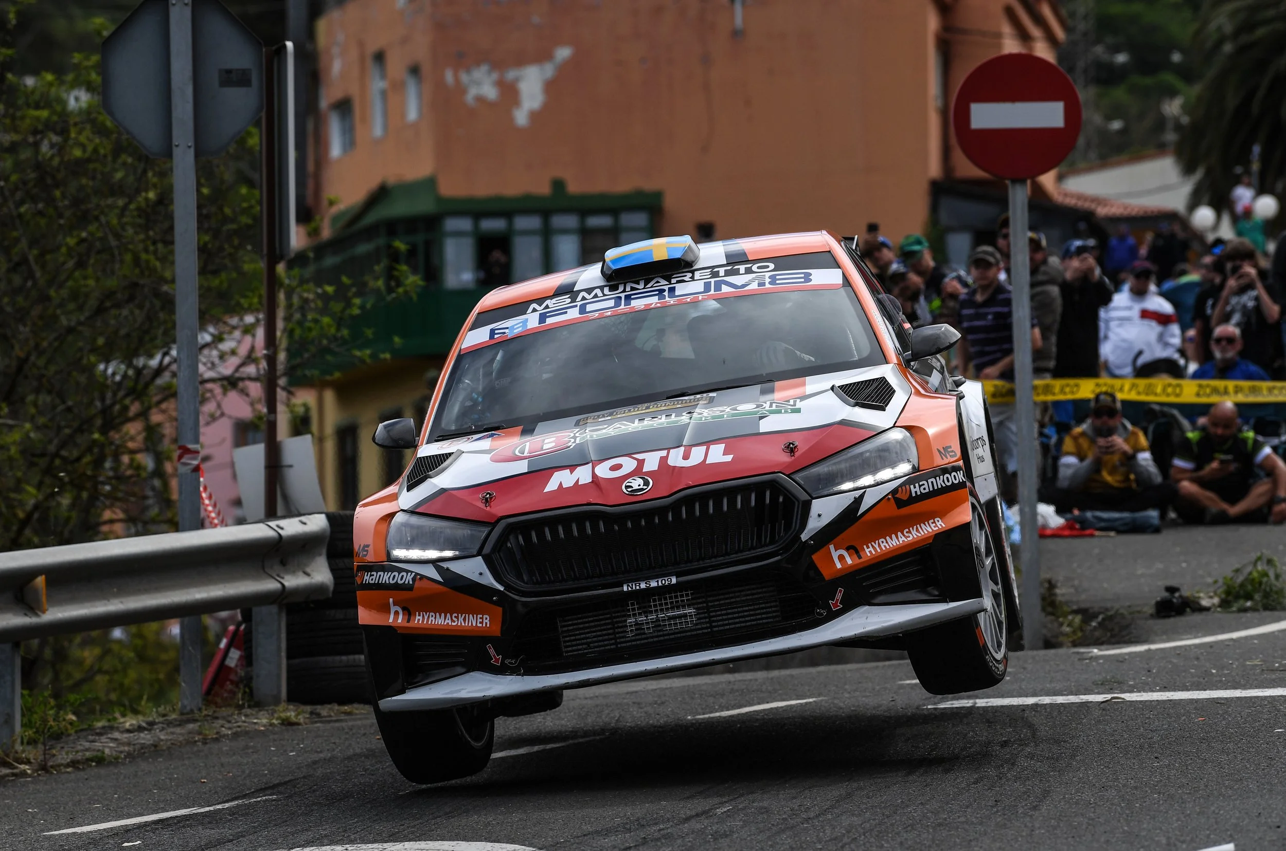 A rally car is in mid-air after going over a bump or jump on a road, surrounded by spectators and road signs.