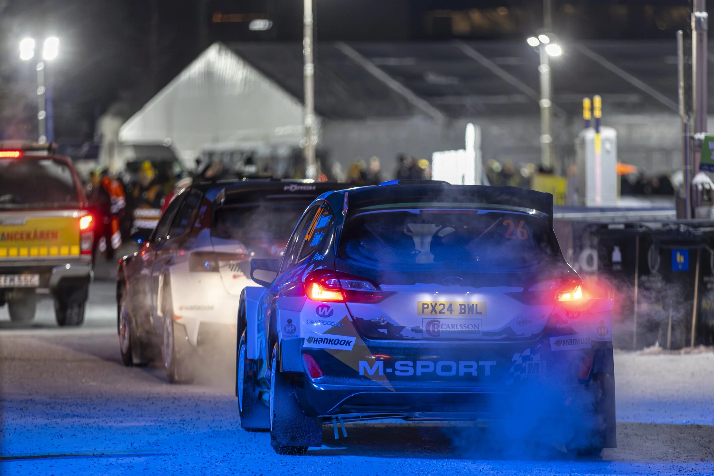Nighttime scene of race cars on a track, with bright lights and a crowd in the background, smoke or dust around the cars, the prominent car in the foreground is a blue and black racing car with the number 26 and various sponsor logos.