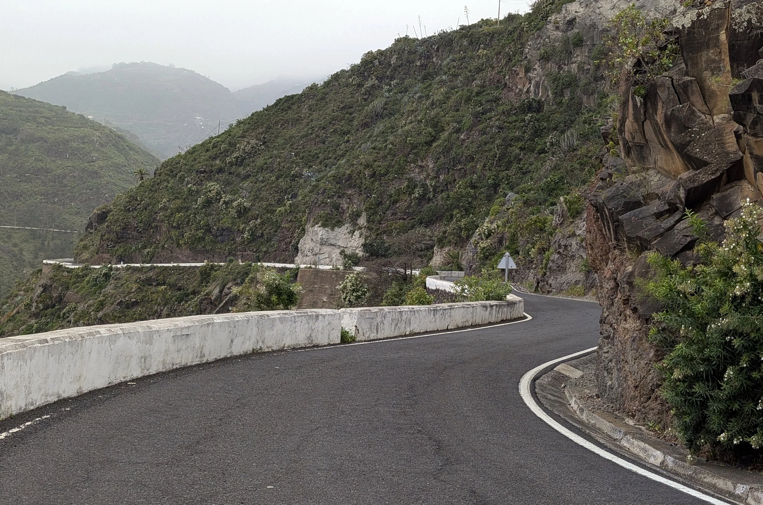 A winding mountain road with a white concrete barrier on one side, surrounded by rocky cliffs, green vegetation, and mountains in the background under cloudy weather.