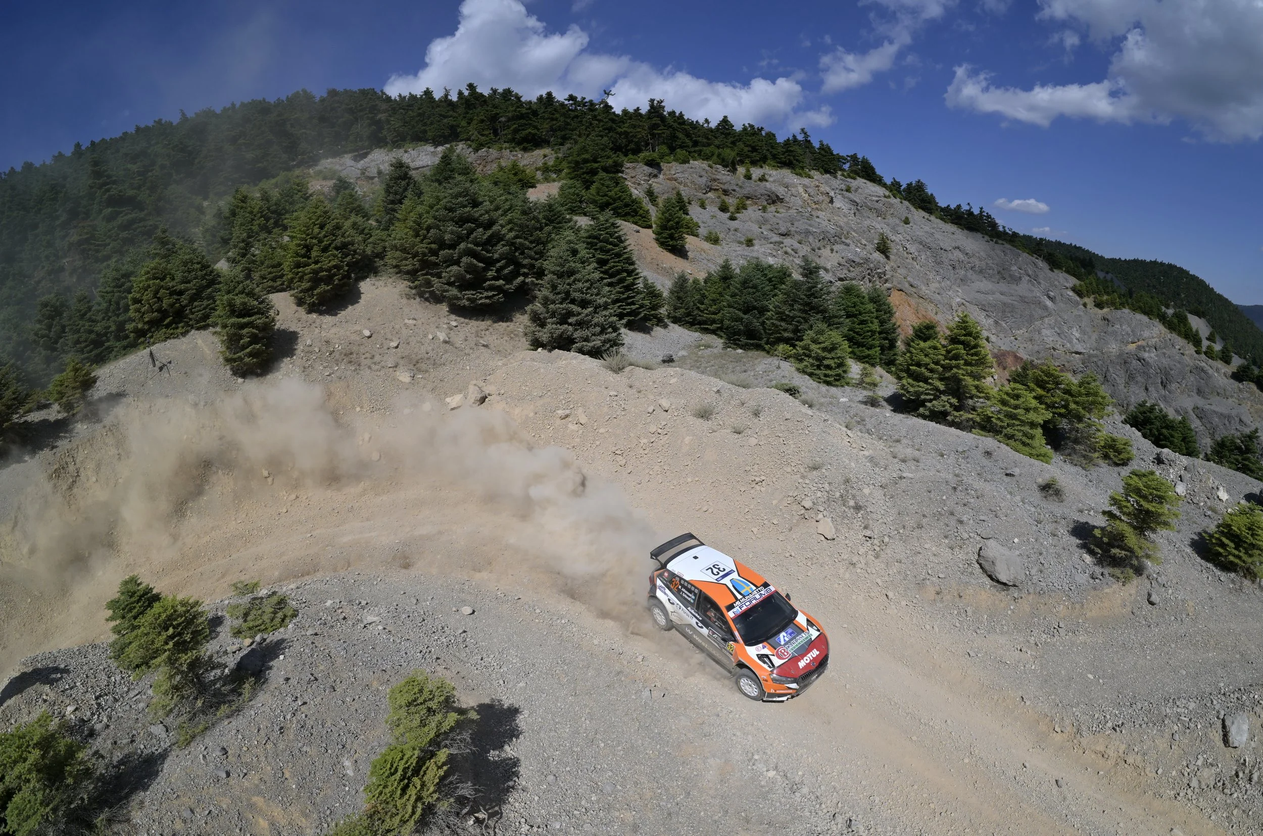 Rally car racing on dirt road by forested mountain with dust trail in the air.