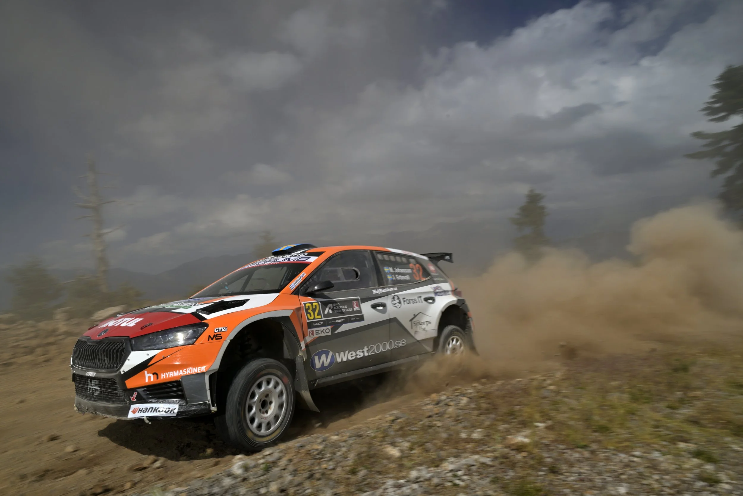 A rally car speeding on a dirt road, kicking up dust, under a cloudy sky with mountains and trees in the background.