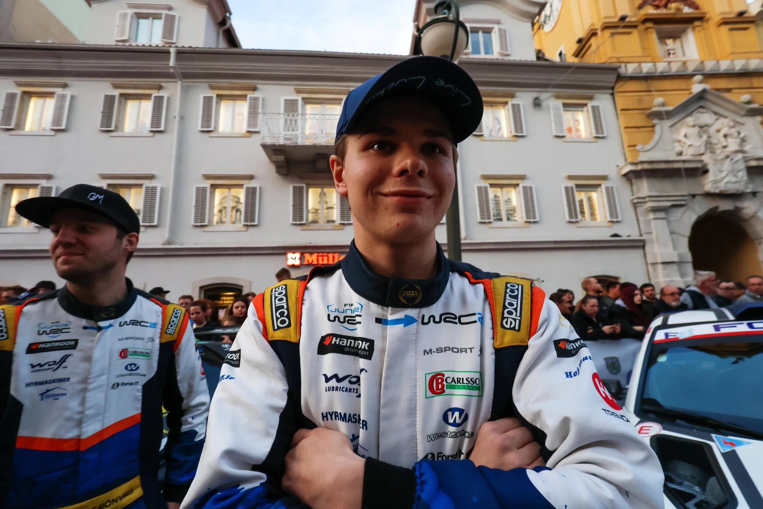 Young male race car driver in racing suit and cap, standing with arms crossed, in front of a crowd and historic building with windows and decorative architecture.