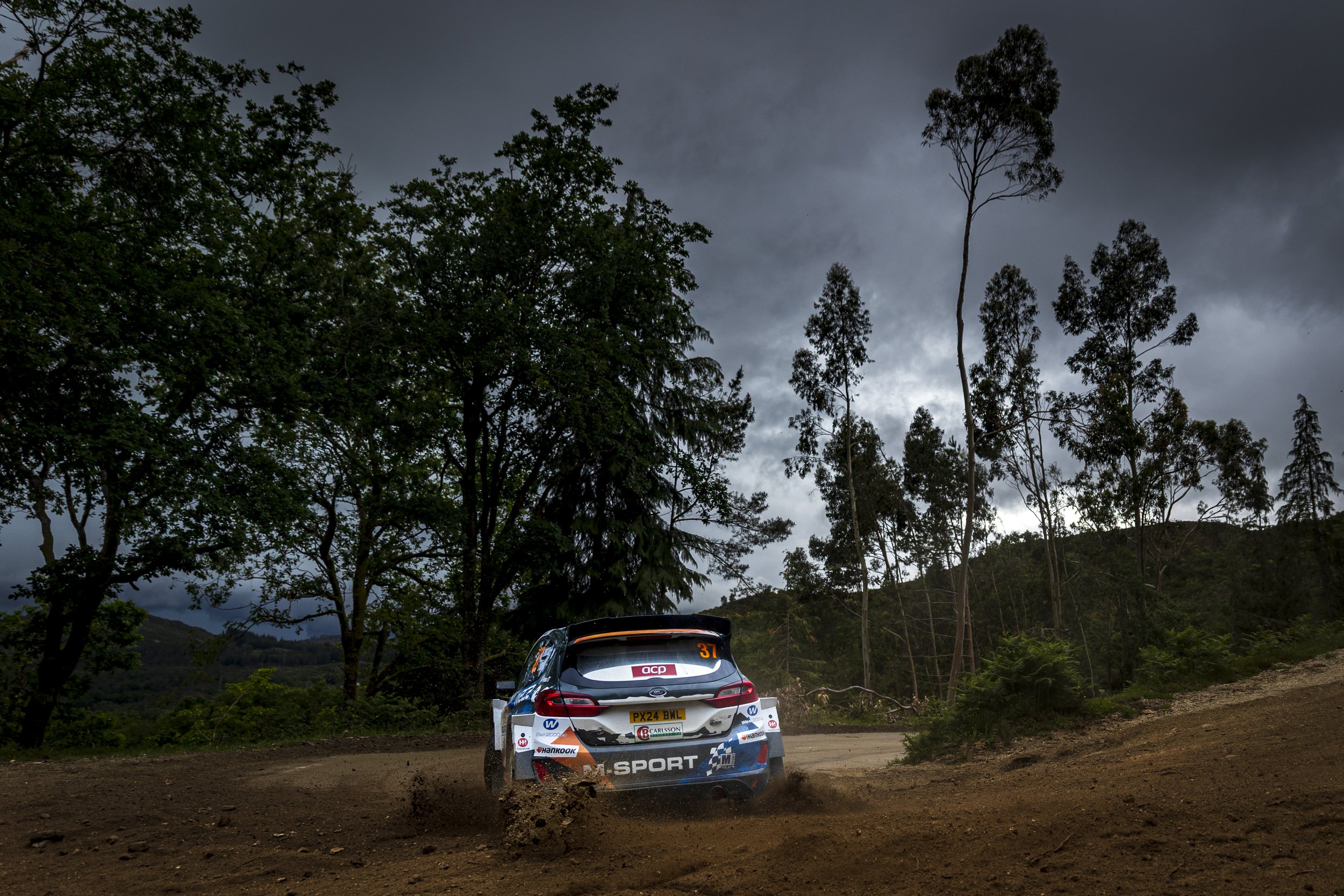 Rally car on dirt road with dust clouds, surrounded by trees and dark cloudy sky.