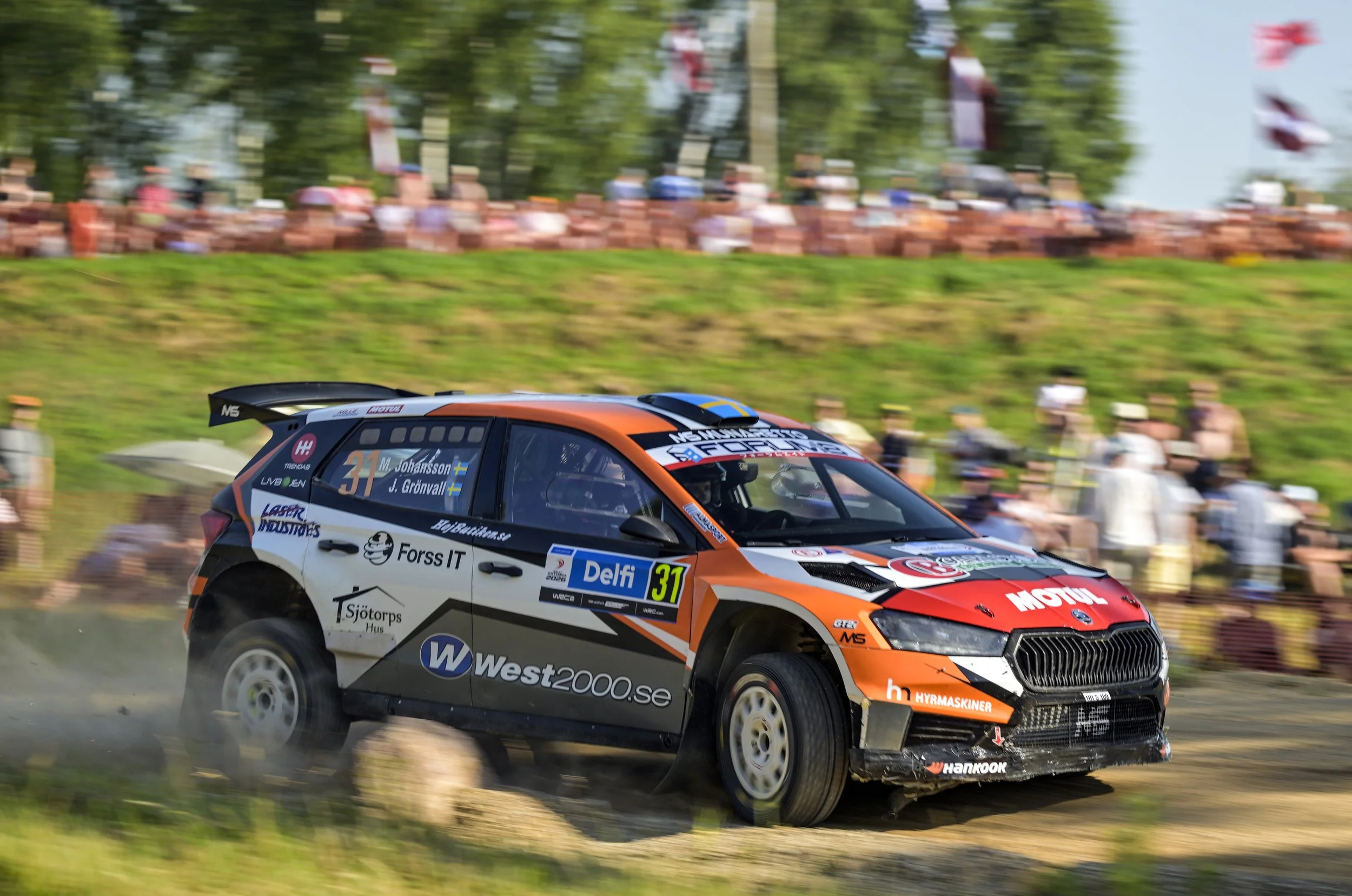 A rally car with the number 31 speeding on a dirt track, kicking up dust, with a crowd of spectators blurred in the background.