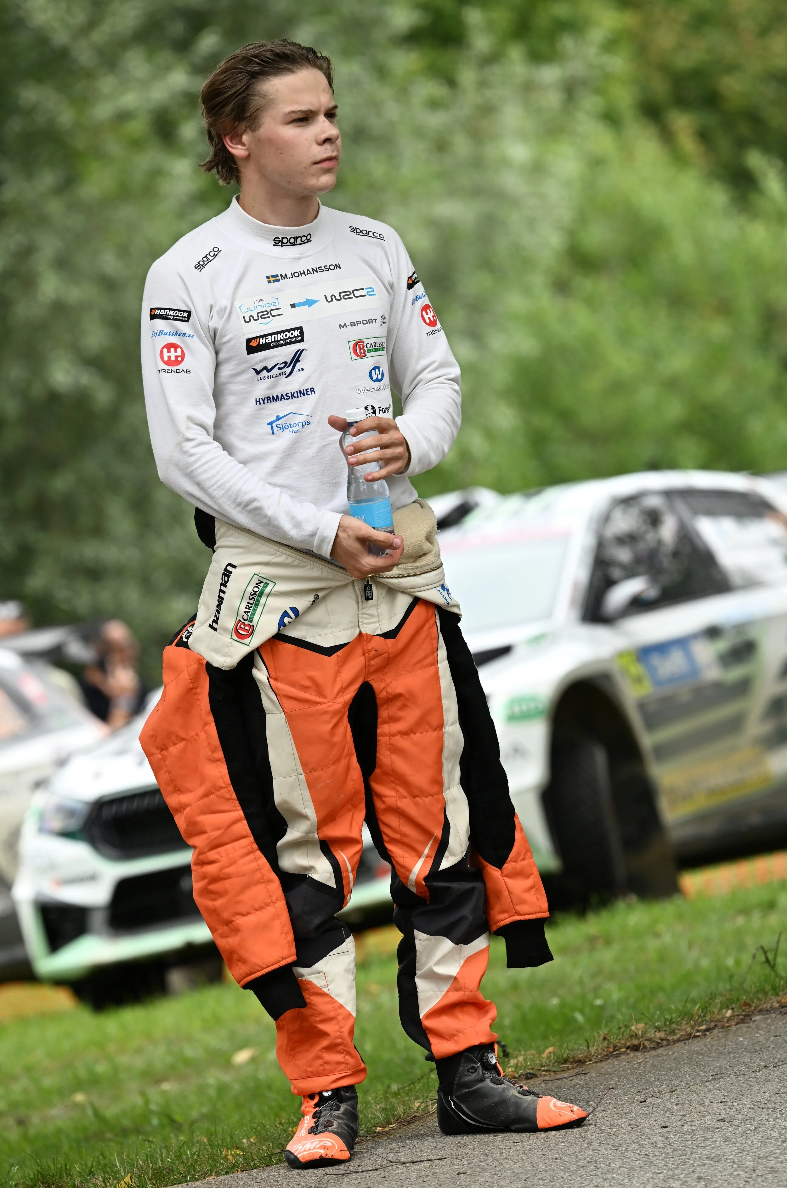 A young rally driver stands outdoors on a grassy area, holding a water bottle, wearing a racing suit with various sponsor logos, and a pair of specialized racing shoes, with rally cars and trees in the background.
