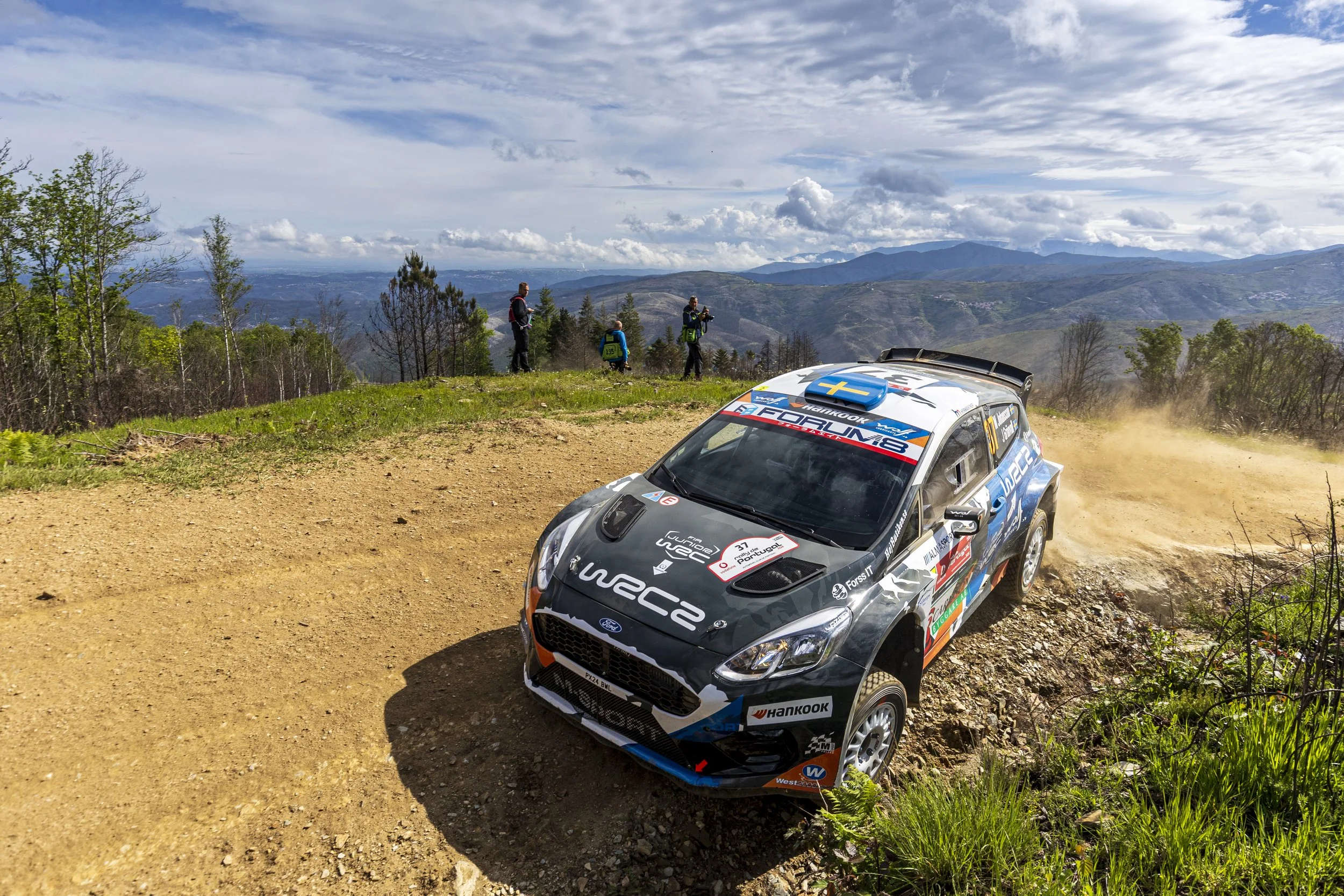 Rally car speeding on a dirt mountain trail with spectators and scenic mountainous landscape in the background.