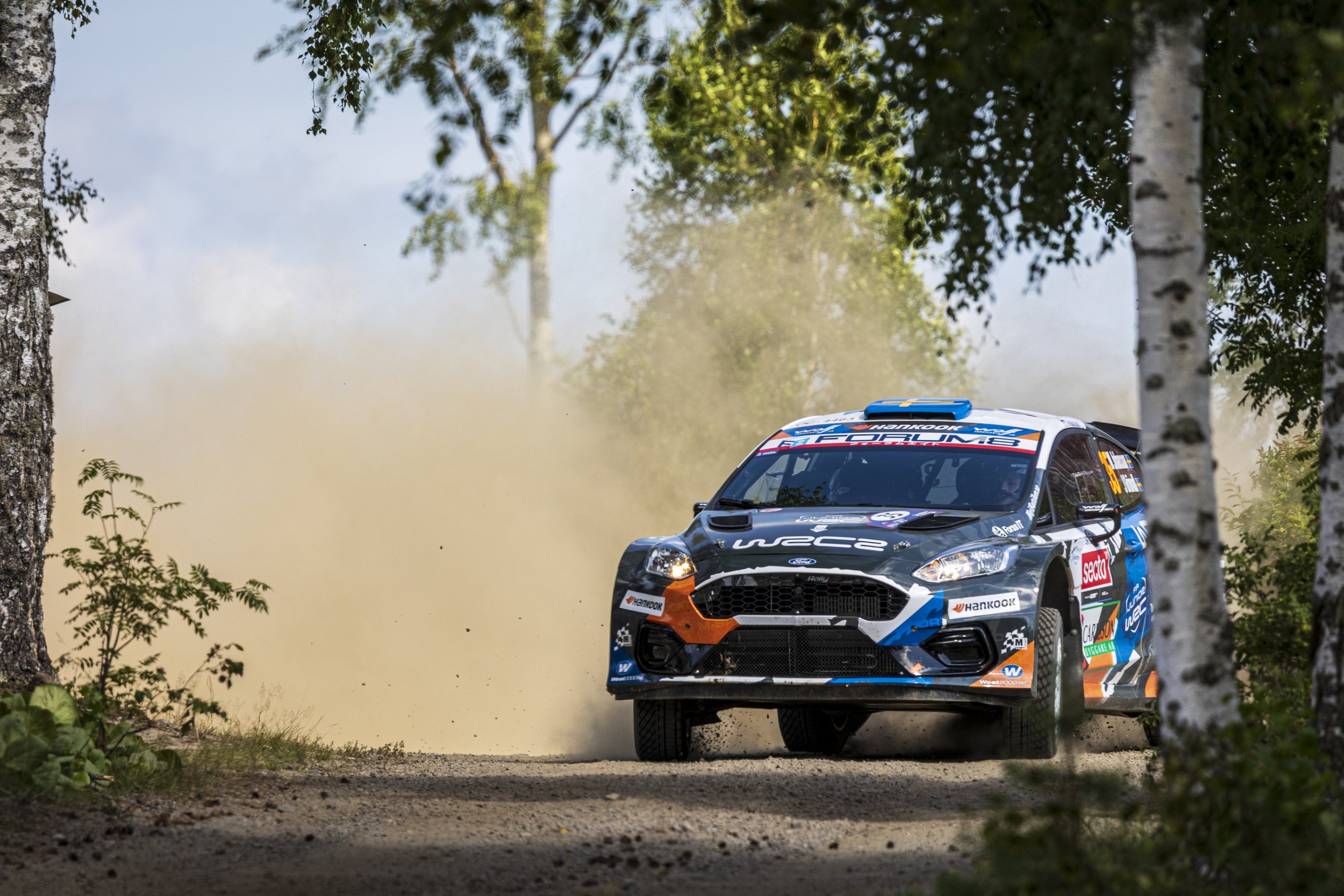 A blue and white rally car kicking up dust as it drives on a dirt road through a wooded area with trees and foliage.