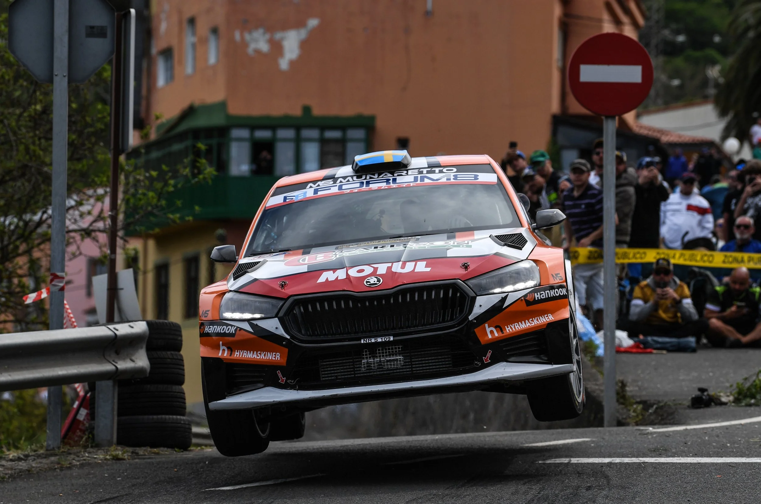A rally car in mid-air during a race, with spectators in the background watching from behind barriers.