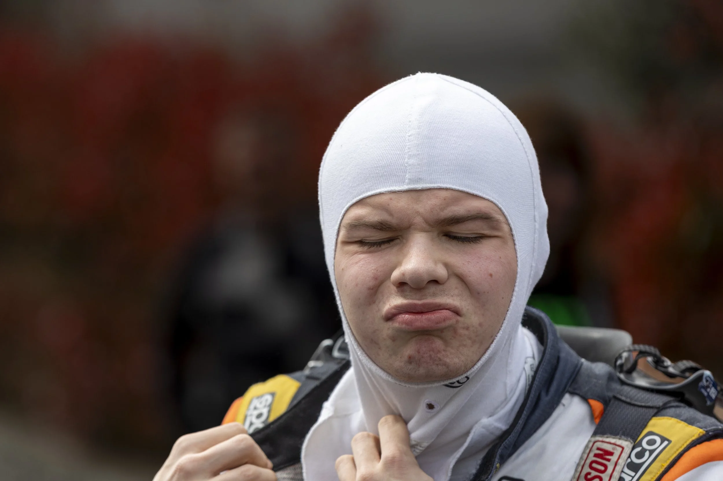 Close-up of a young man with a white racing suit and a white cap, with his eyes closed and a serious expression, outdoors.