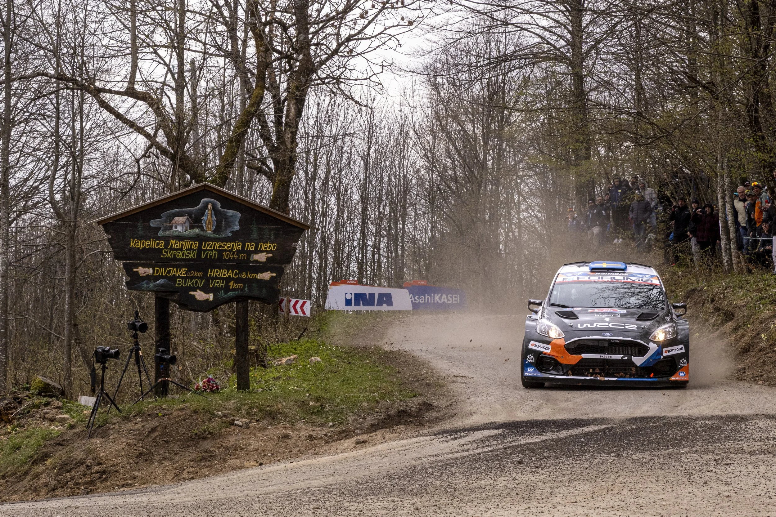 Rally car driving on a dirt road in a wooded area with spectators watching from the side.