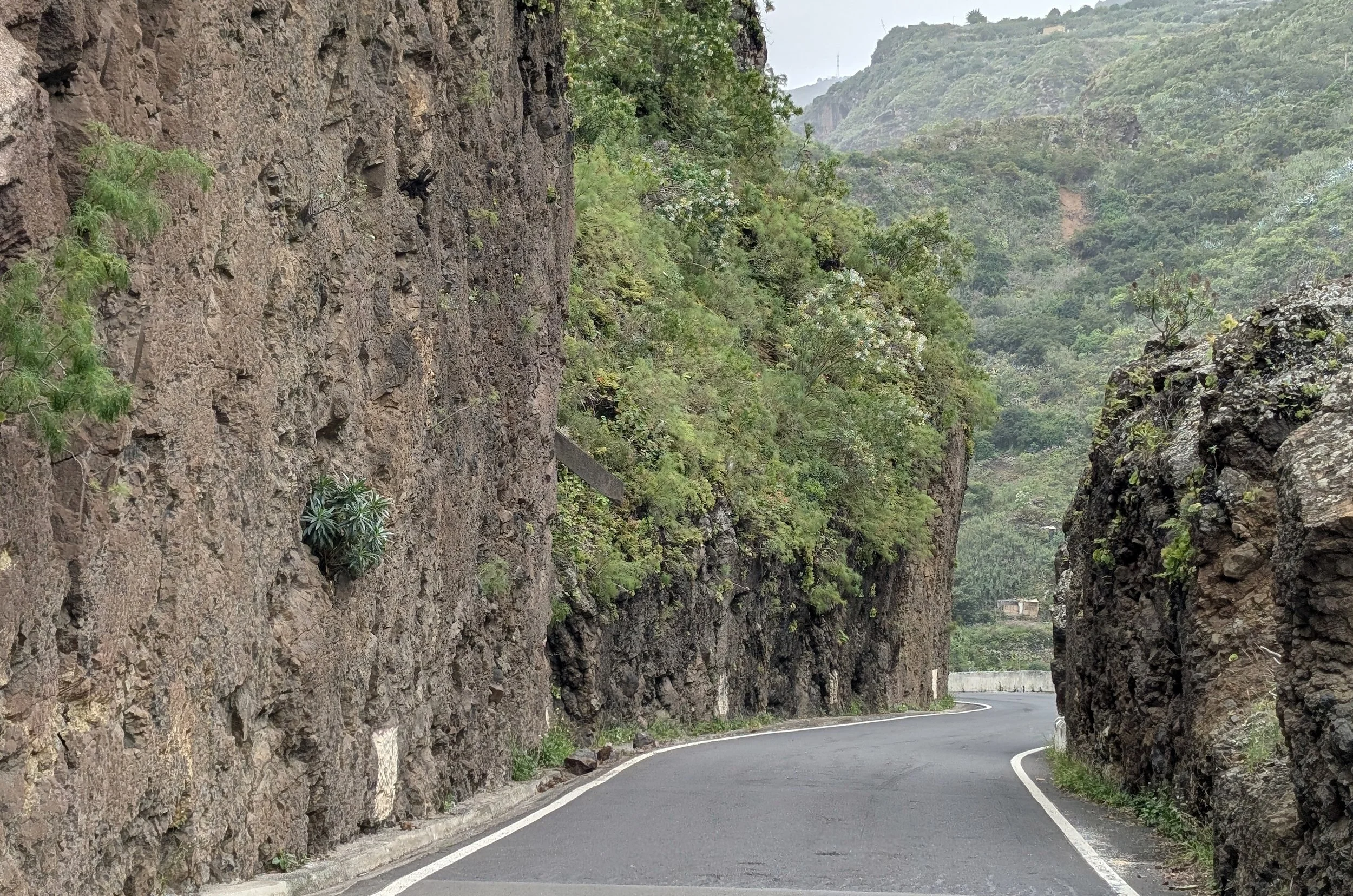 A winding mountain road passing through a narrow rocky canyon with steep cliff walls on both sides, and lush green hills in the background.