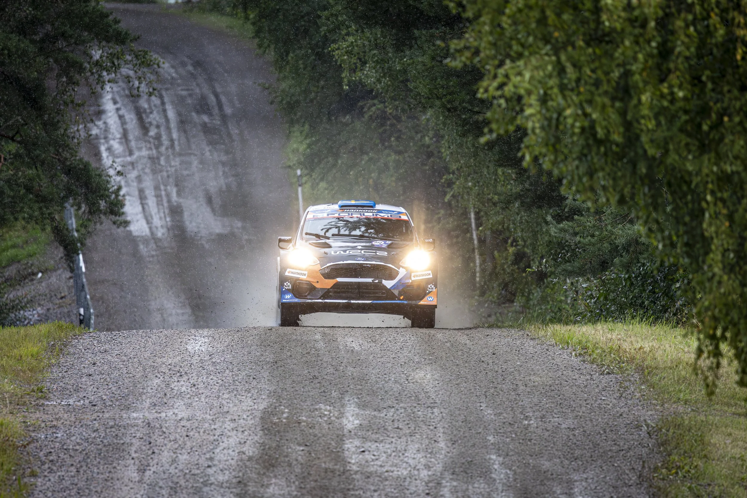 A rally car driving on a gravel road through a wooded area, splashing water and mud as headlights shine, surrounded by green trees and bushes.