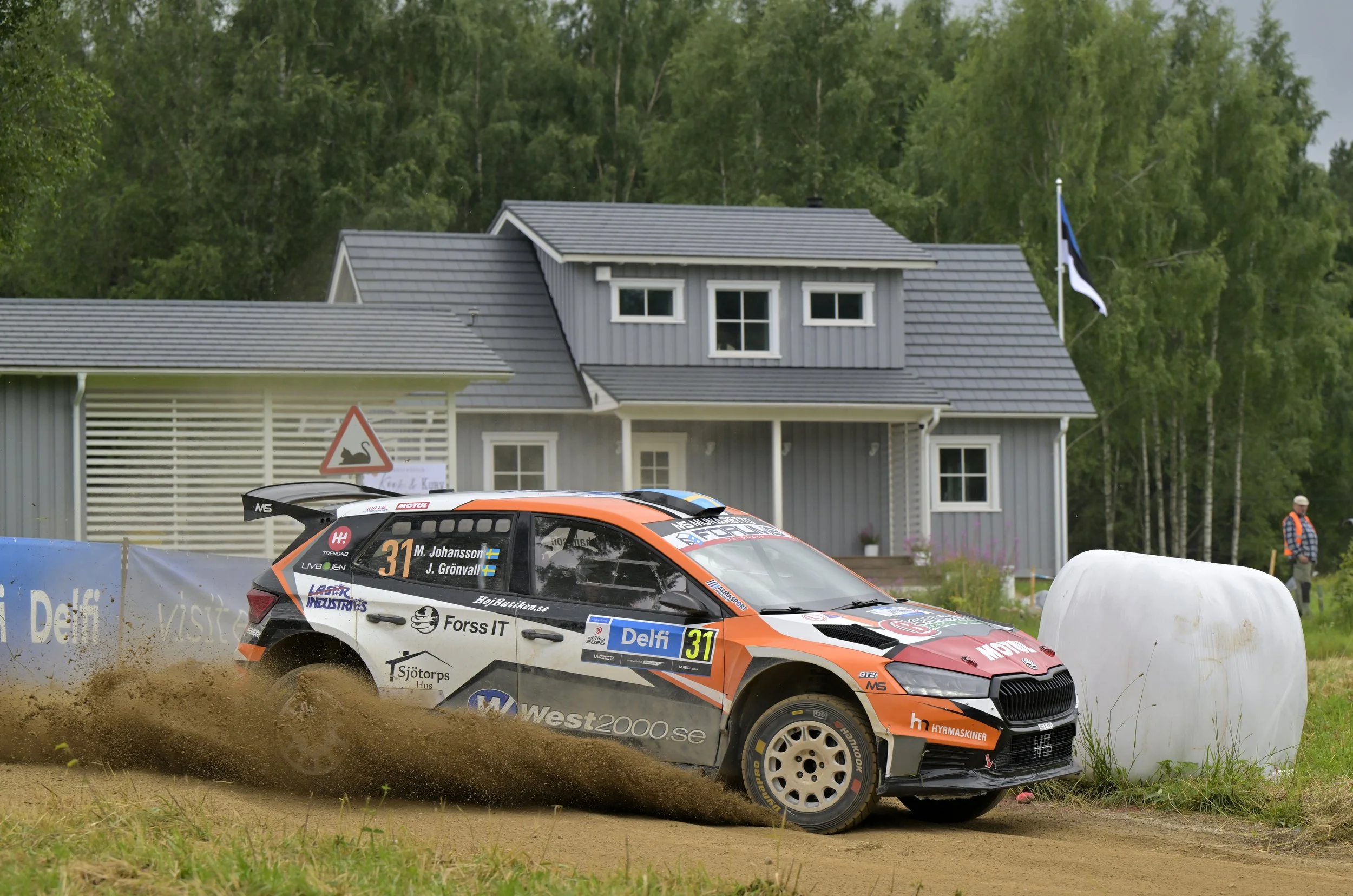 A rally car with the number 31 on its side driving through a dirt track, kicking up dust, during a race with a house and trees in the background.