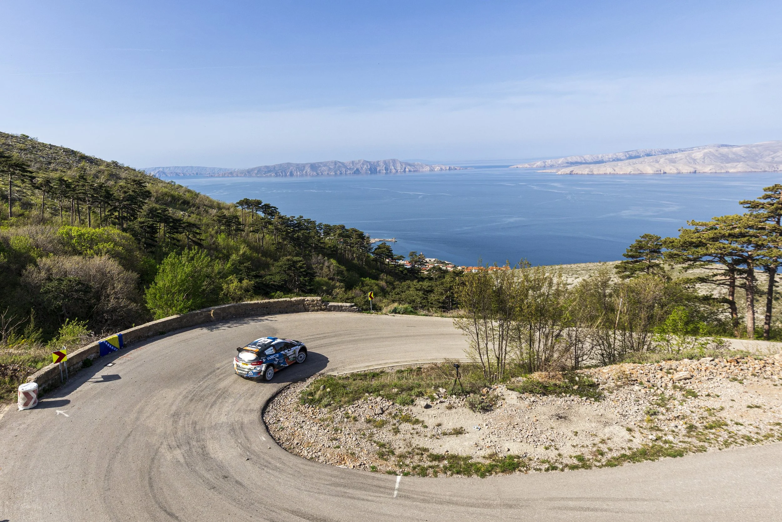A rally car speeding around a sharp bend on a mountain road overlooking a large body of water with mountains in the distance. The road is narrow with barriers and caution signs.