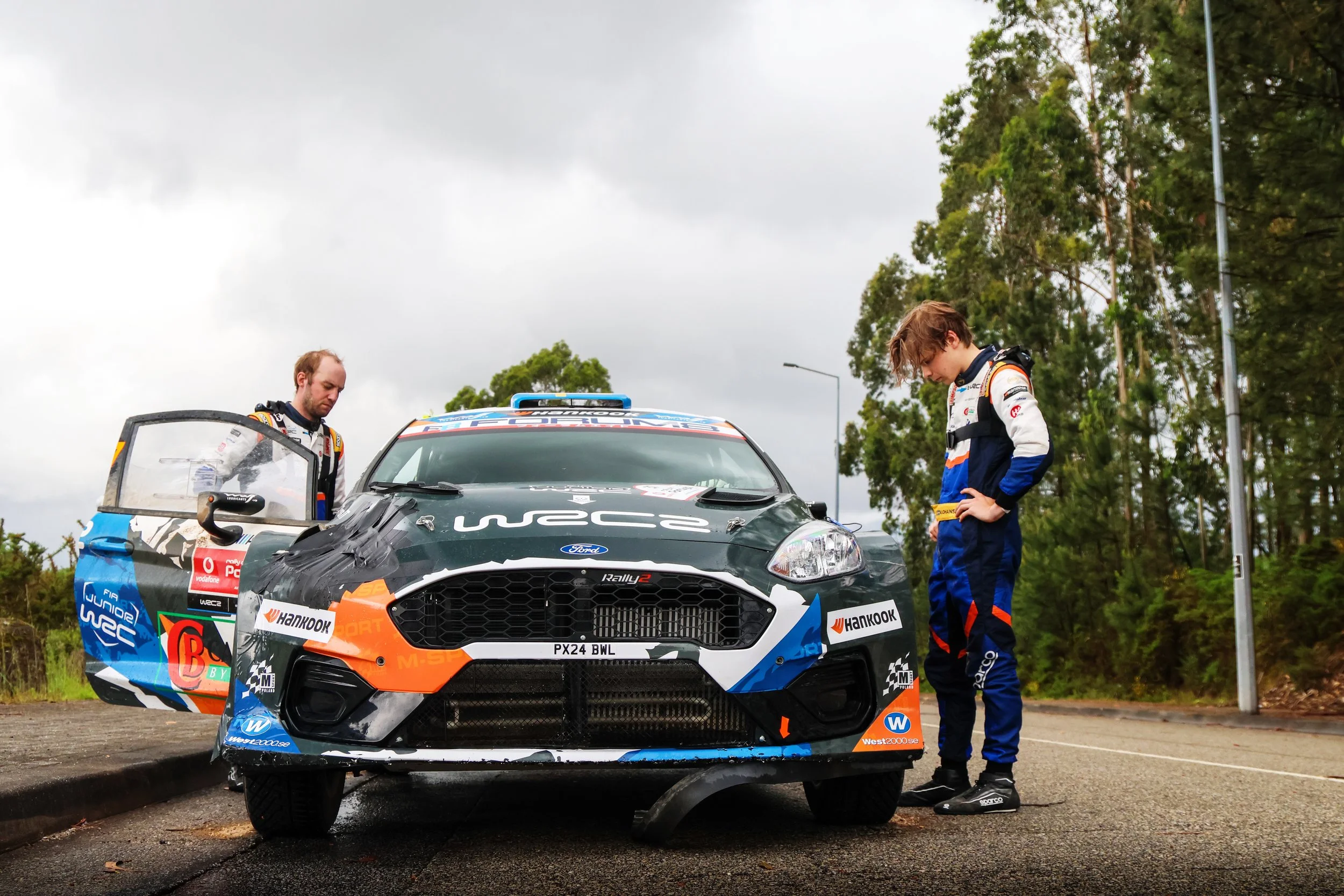 Two young male rally drivers in racing suits standing near a rally car, one looking down and the other with hands on hips, with trees and cloudy sky in the background.