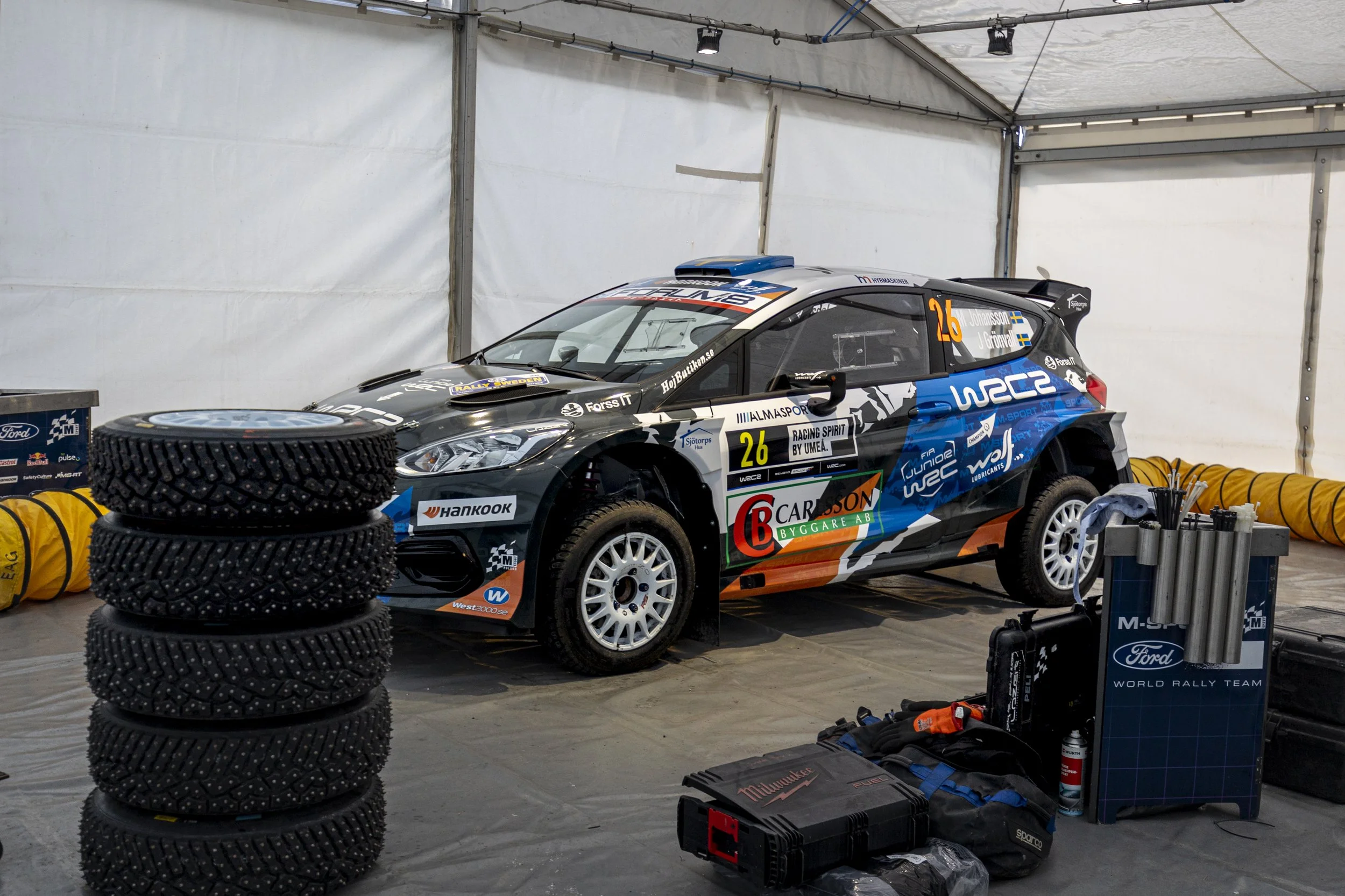 Race car inside a garage with tires, tools, and equipment around it, marked with number 26 and various sponsor logos.
