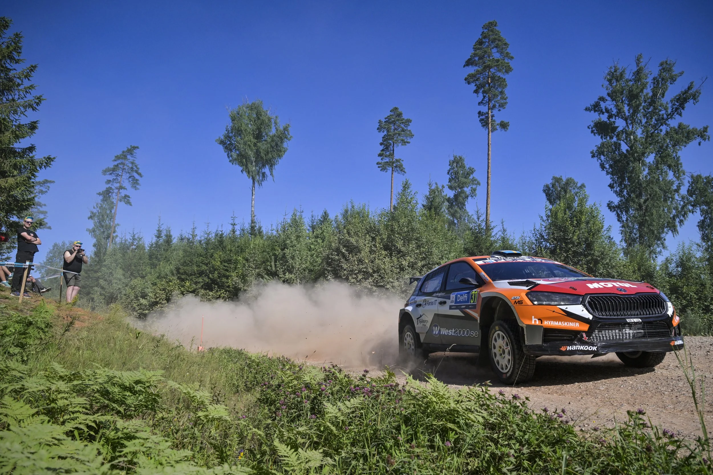 A rally car racing on a dirt trail in a forested area with spectators watching. The sky is clear and blue, with tall trees in the background.