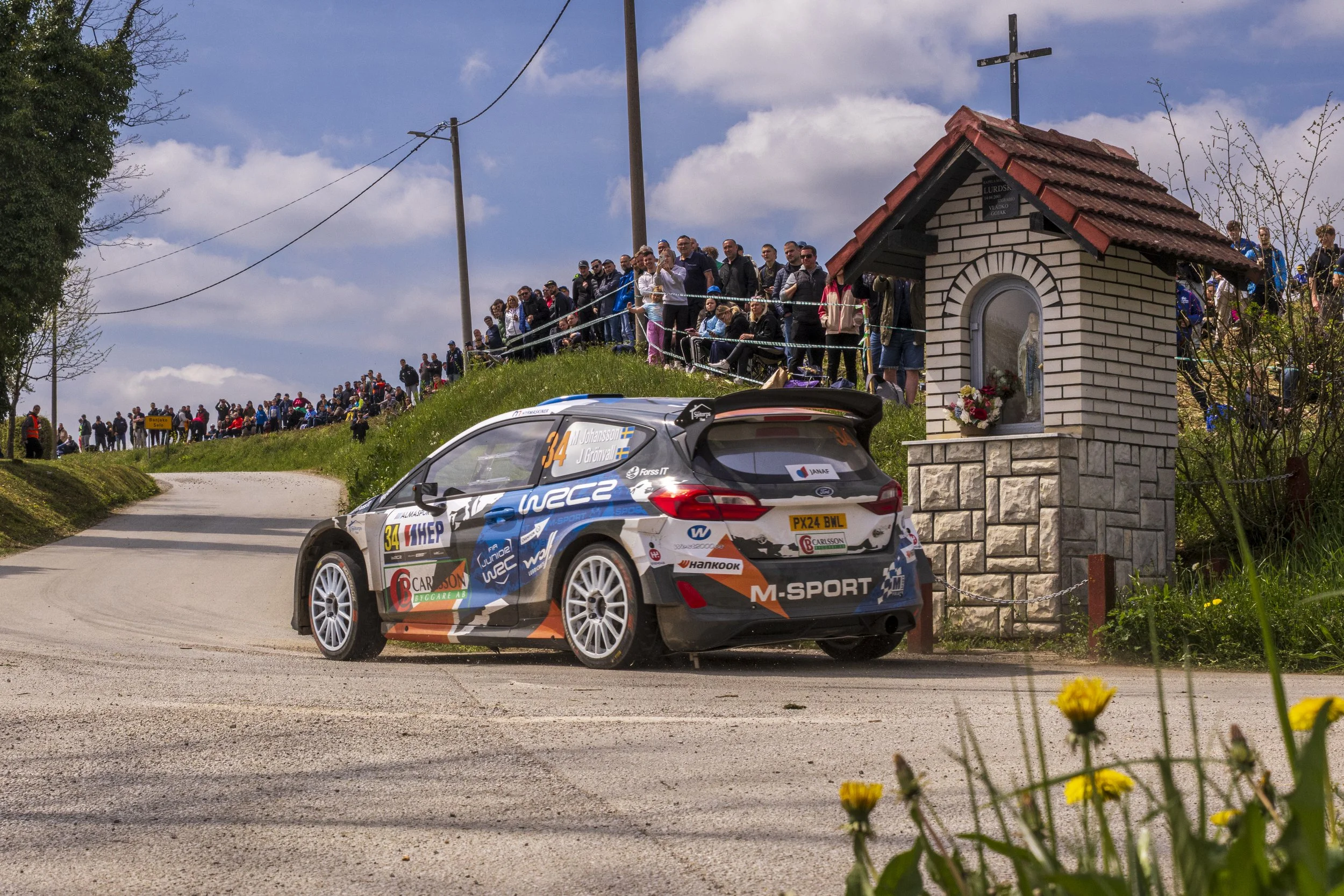 A rally car speeds along a rural dirt road with a crowd of spectators watching from a grassy hill. The car is decorated with numerous sponsor logos and the numbers 34 and features a Swedish flag. Nearby, there is a small brick shrine with a cross on 