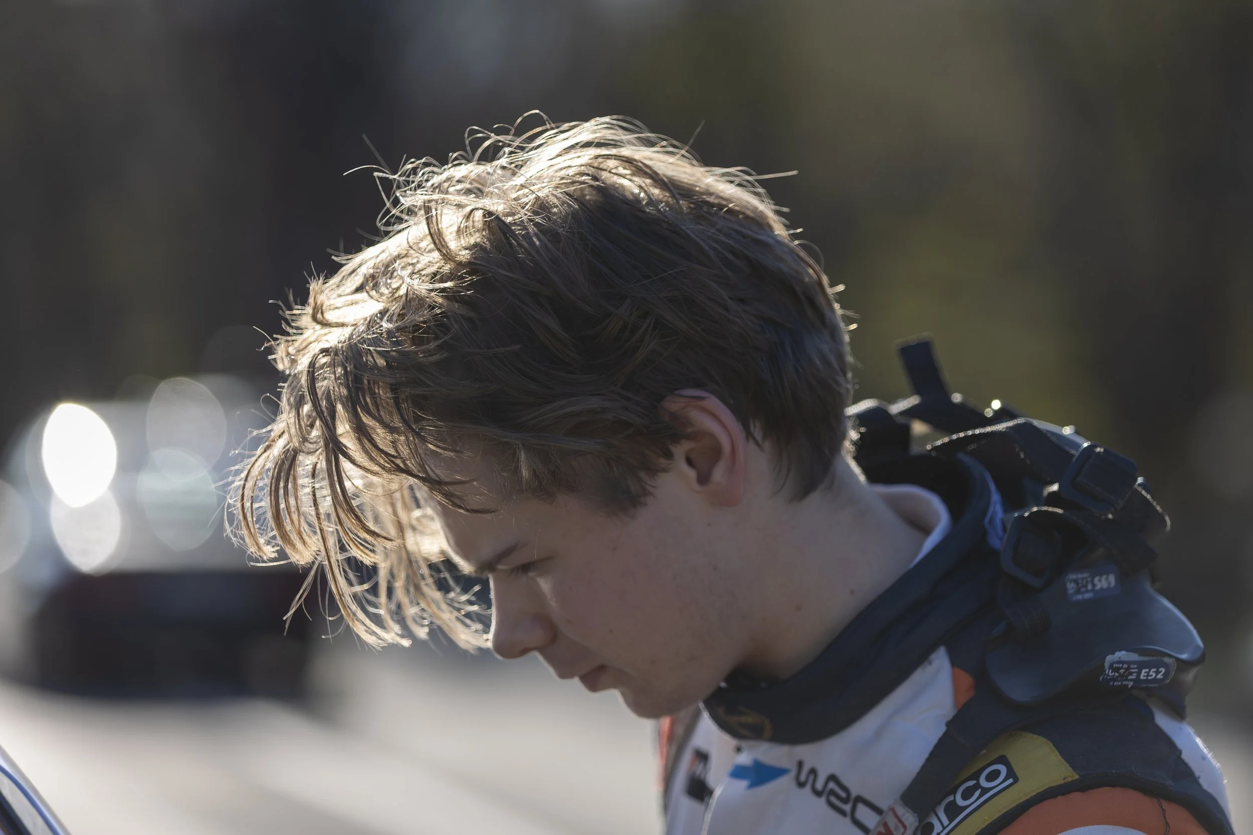 Close-up of a young male cyclist with messy blond hair, wearing a racing jersey and a harness strap, looking downward with a serious expression, illuminated by sunlight.