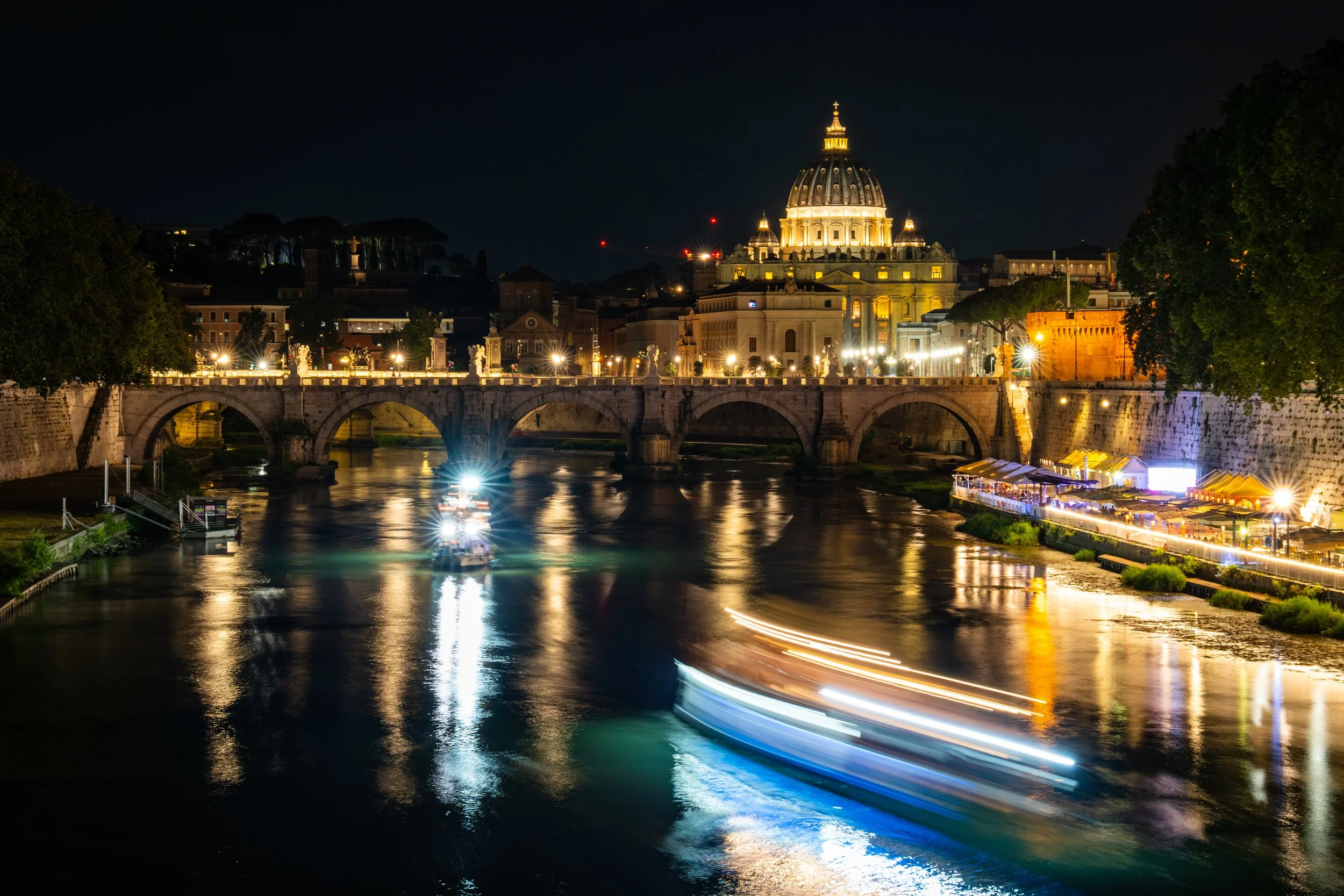 Nighttime view of St. Peter's Basilica in Vatican City illuminated, with a bridge over the Tiber River and city lights reflecting on the water.