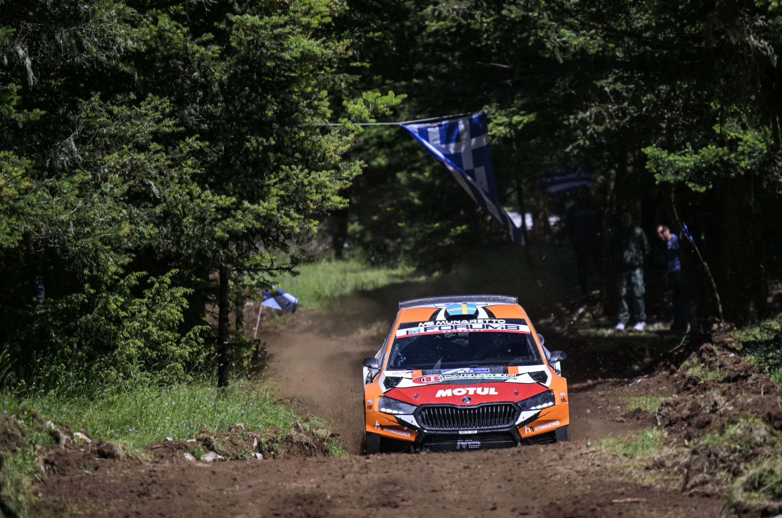 An orange rally car speeding on a dirt road through a forest, with a blue and white checkered flag hanging among green trees in the background.