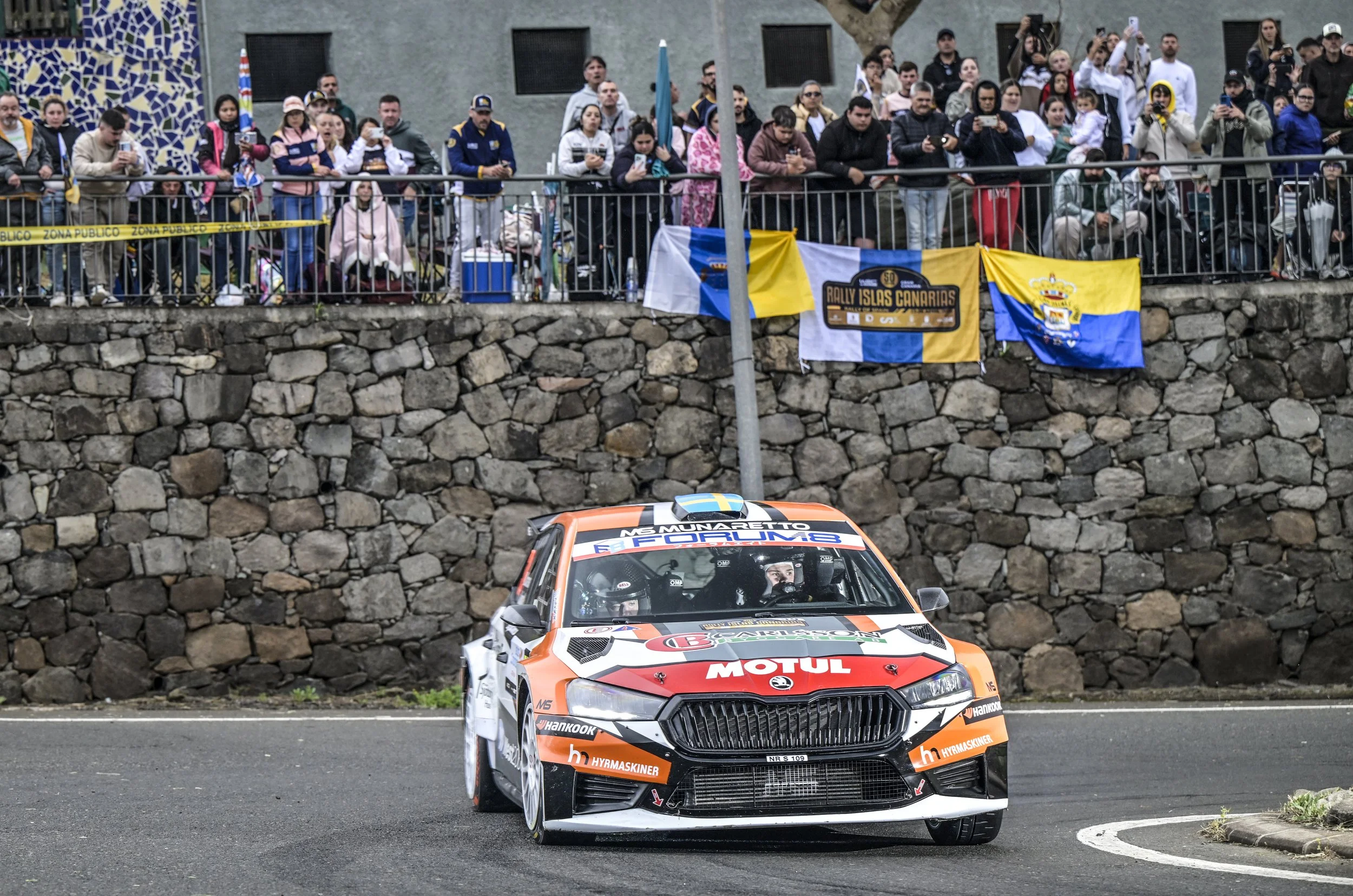 A rally car with a white and orange color scheme racing on a street, with spectators standing behind a railing, some taking photos, and rally flags displayed on the railing.