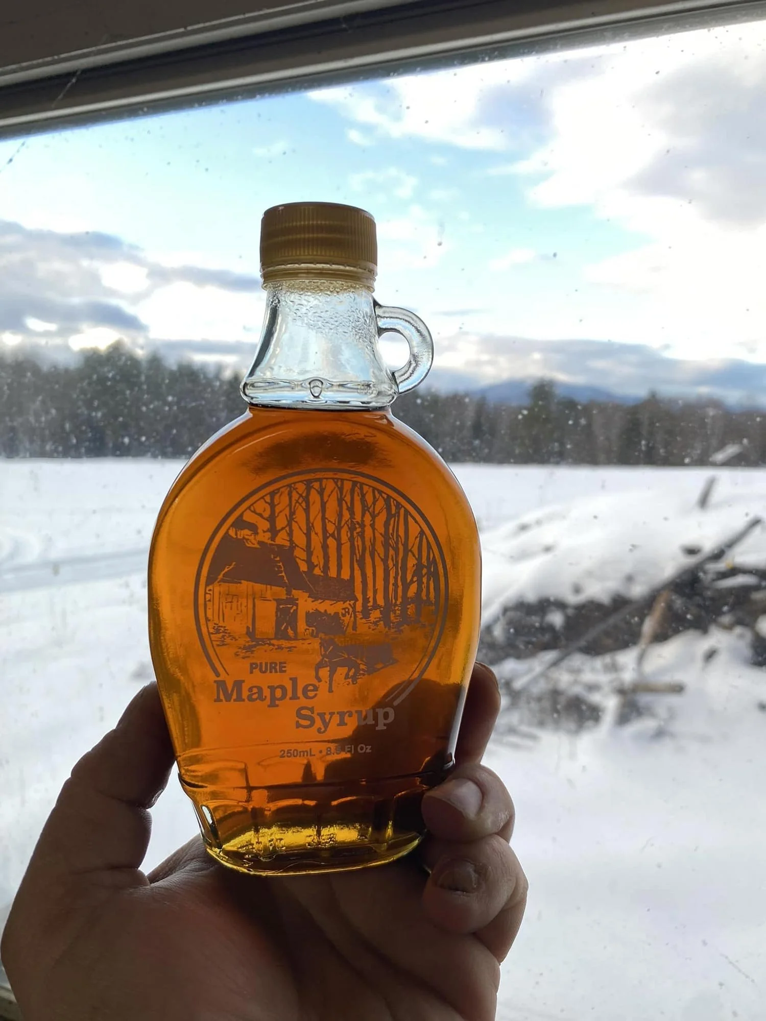 A hand holding a bottle of pure maple syrup near a window with a snowy landscape and cloudy sky outside.