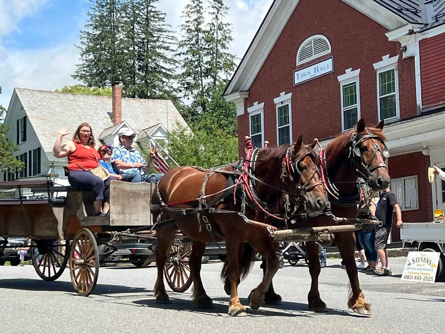 A horse-drawn wagon carrying pasengers, moving through a street in a small town. The background includes a brick town hall building and other buildings, with people walking along the sidewalk and parked cars.