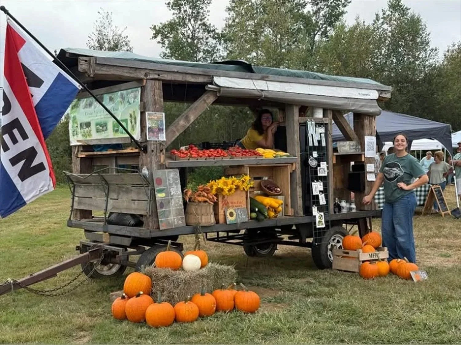 A farm stand with open flag and pumpkins and flowers in front with two women smiling, one standing beside the stand and one sitting inside.