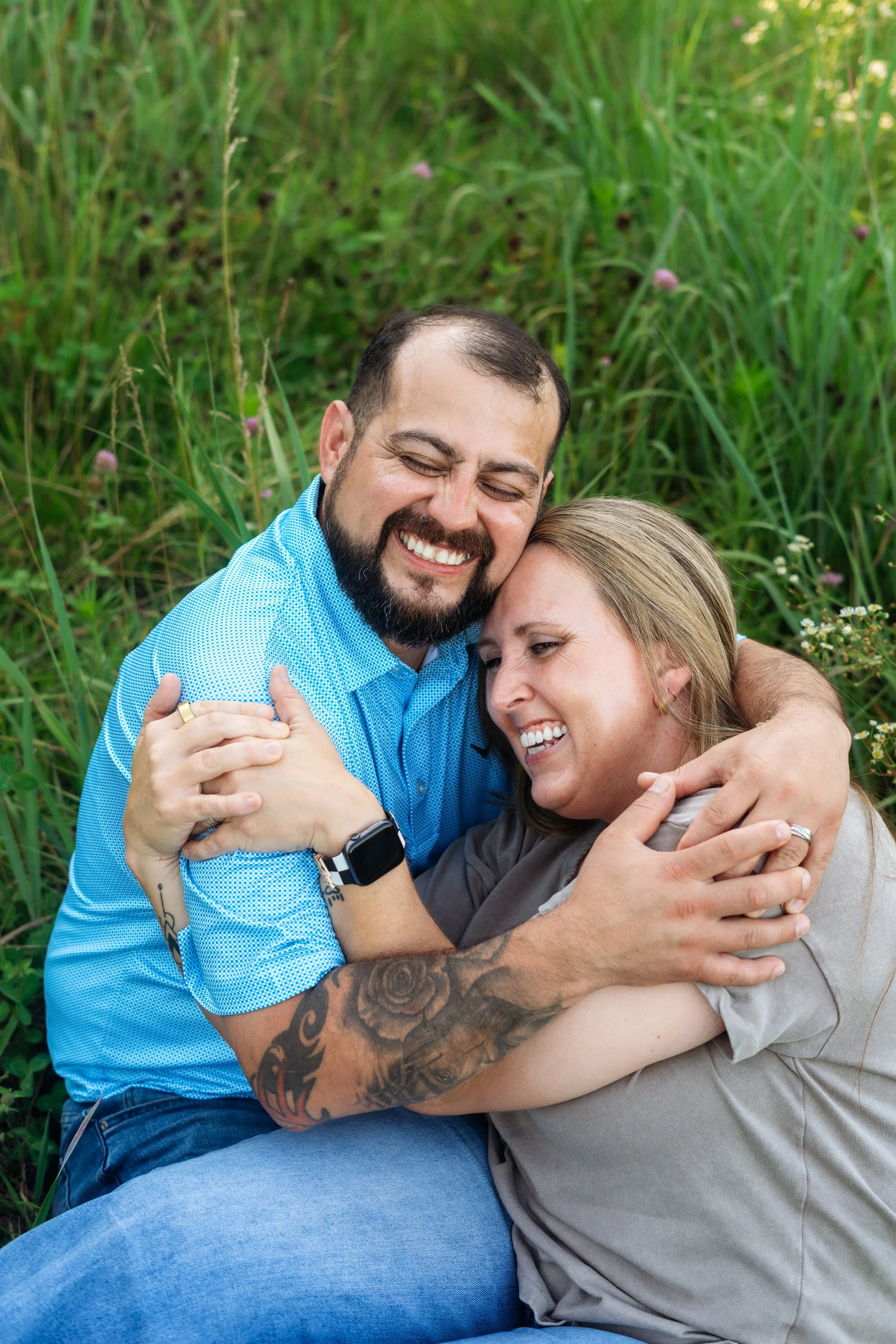 A man with a beard and a woman with blonde hair hugging each other and smiling happily outside in a grassy area with pink and white flowers.