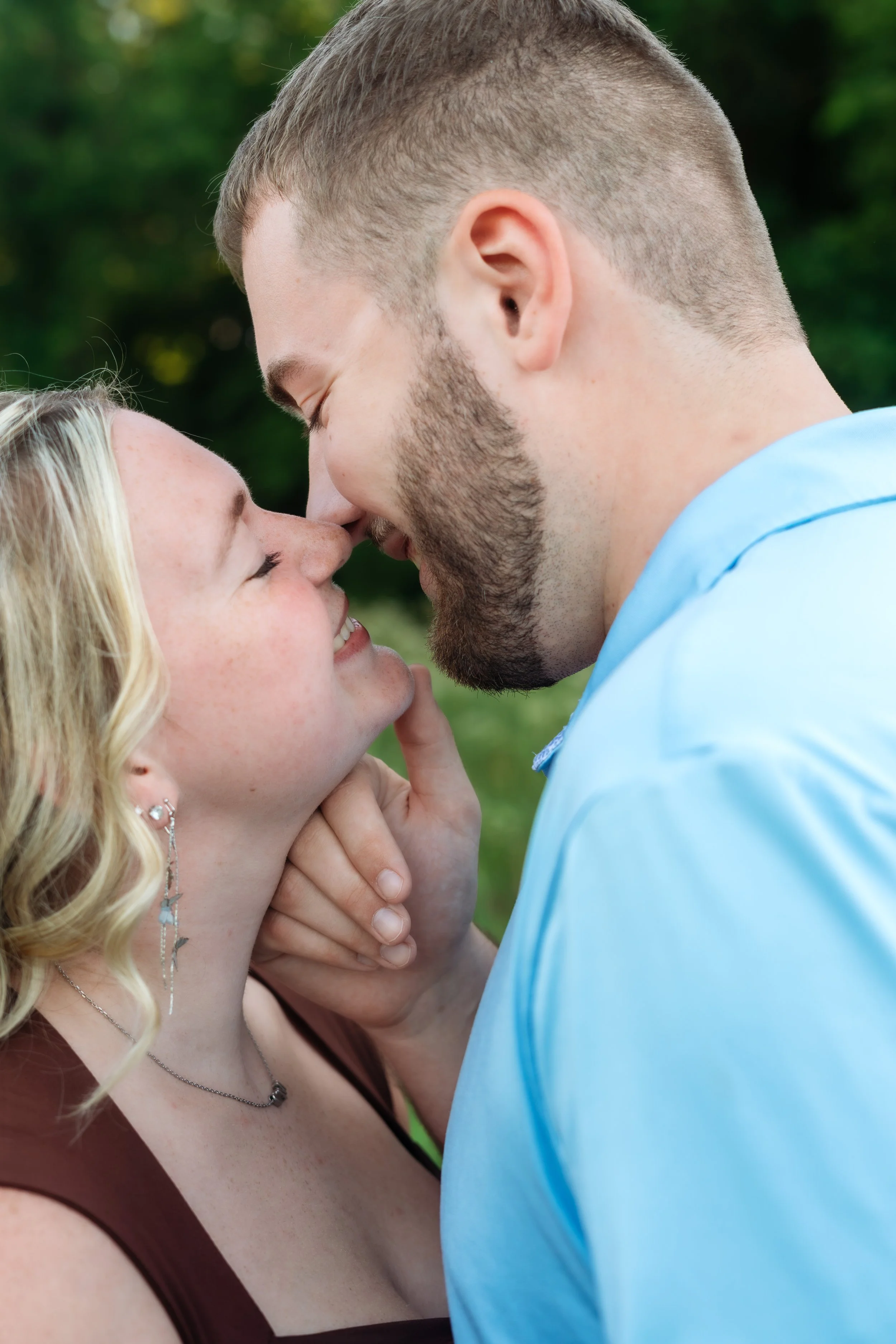 A close-up photo of a couple touching foreheads and smiling outdoors. The woman has wavy blonde hair, earrings, and a necklace, and is wearing a brown top. The man has short hair, a beard, and is wearing a light blue shirt. The background is blurred greenery.