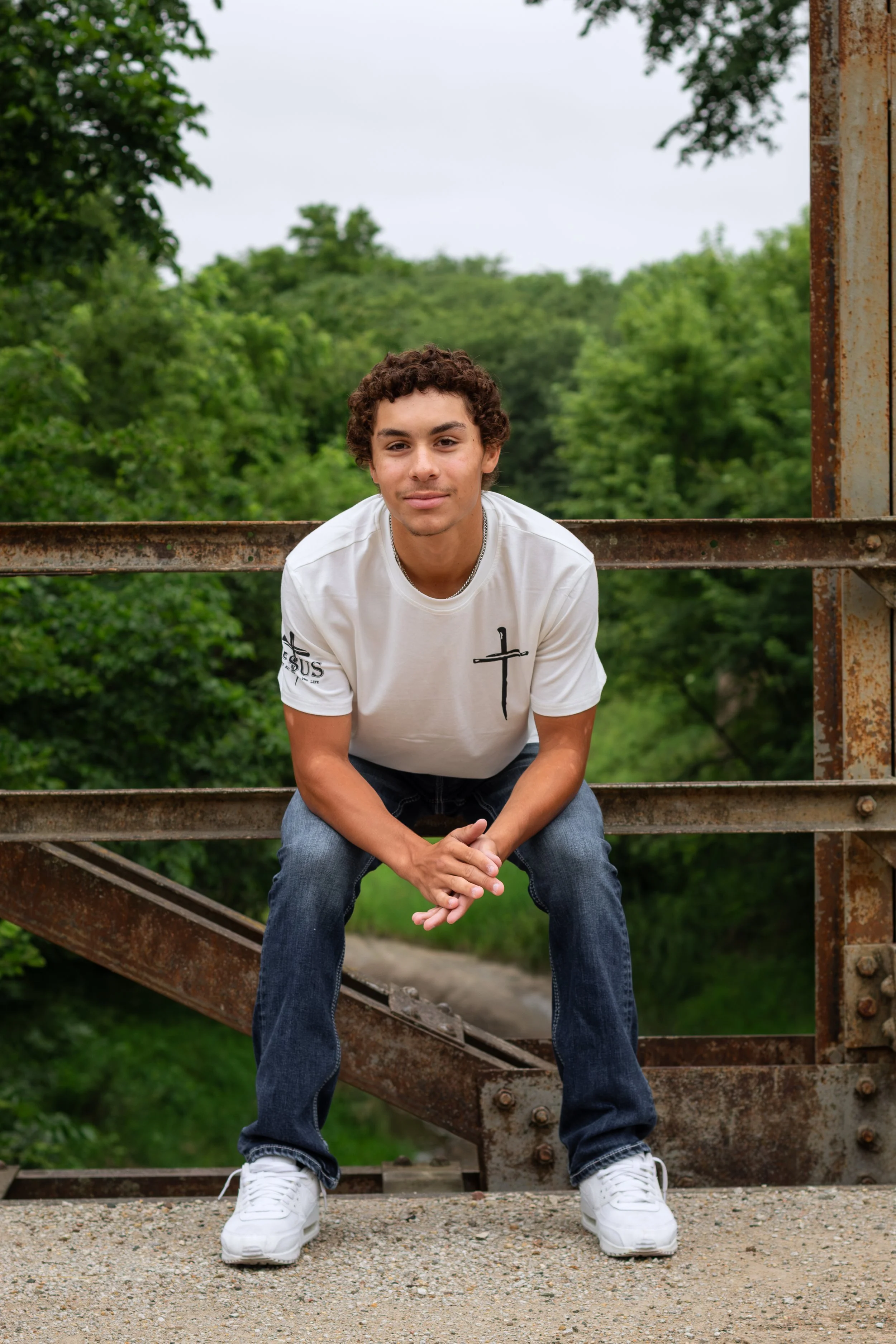 A young man with curly hair sitting on a rusted metal structure outdoors, with a background of green trees and an overcast sky.
