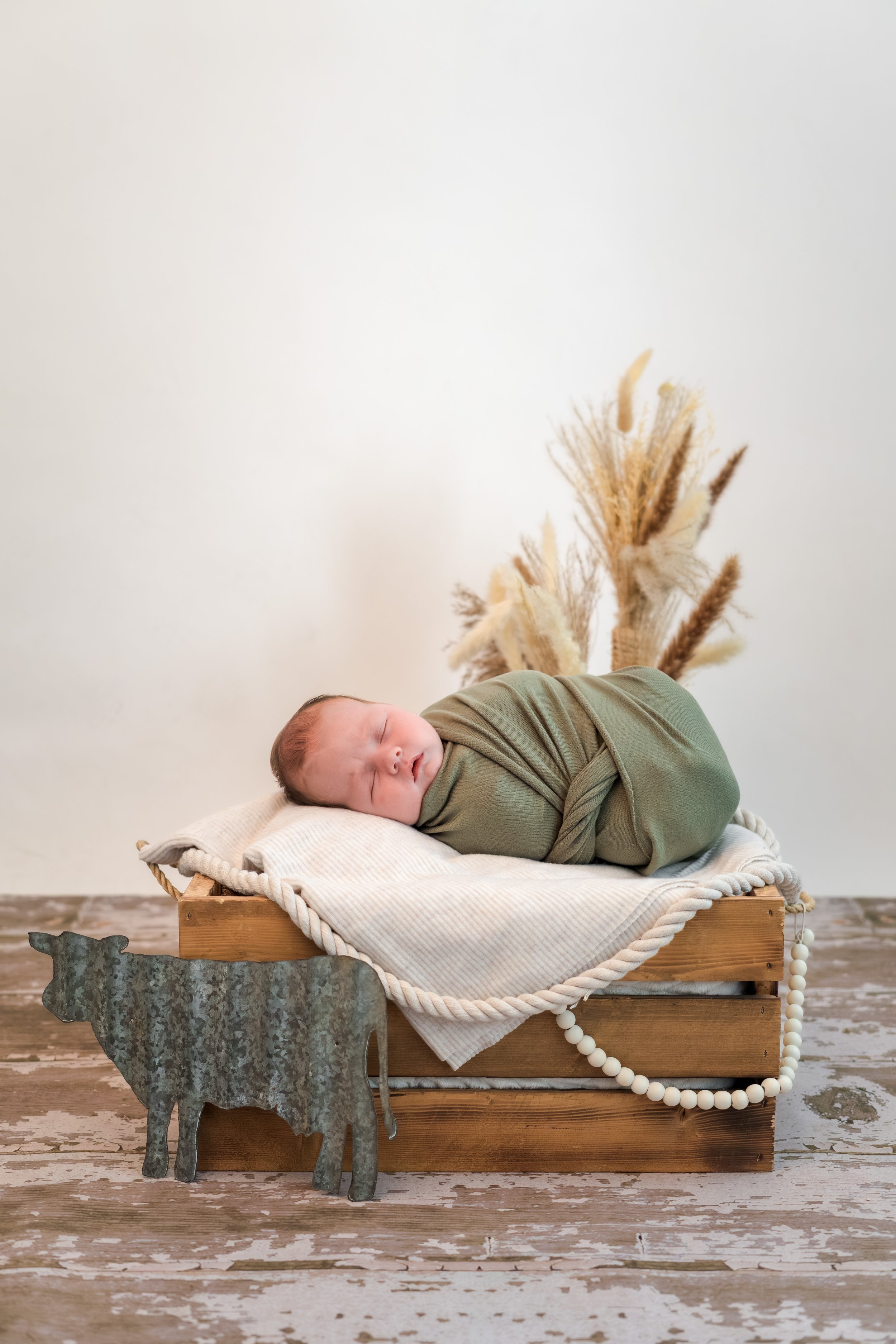 A sleeping baby lying on a small wooden bed with a cream-colored blanket, dressed in a green outfit, on a rustic wooden floor against a plain white wall background with dried pampas grass in a vase behind.