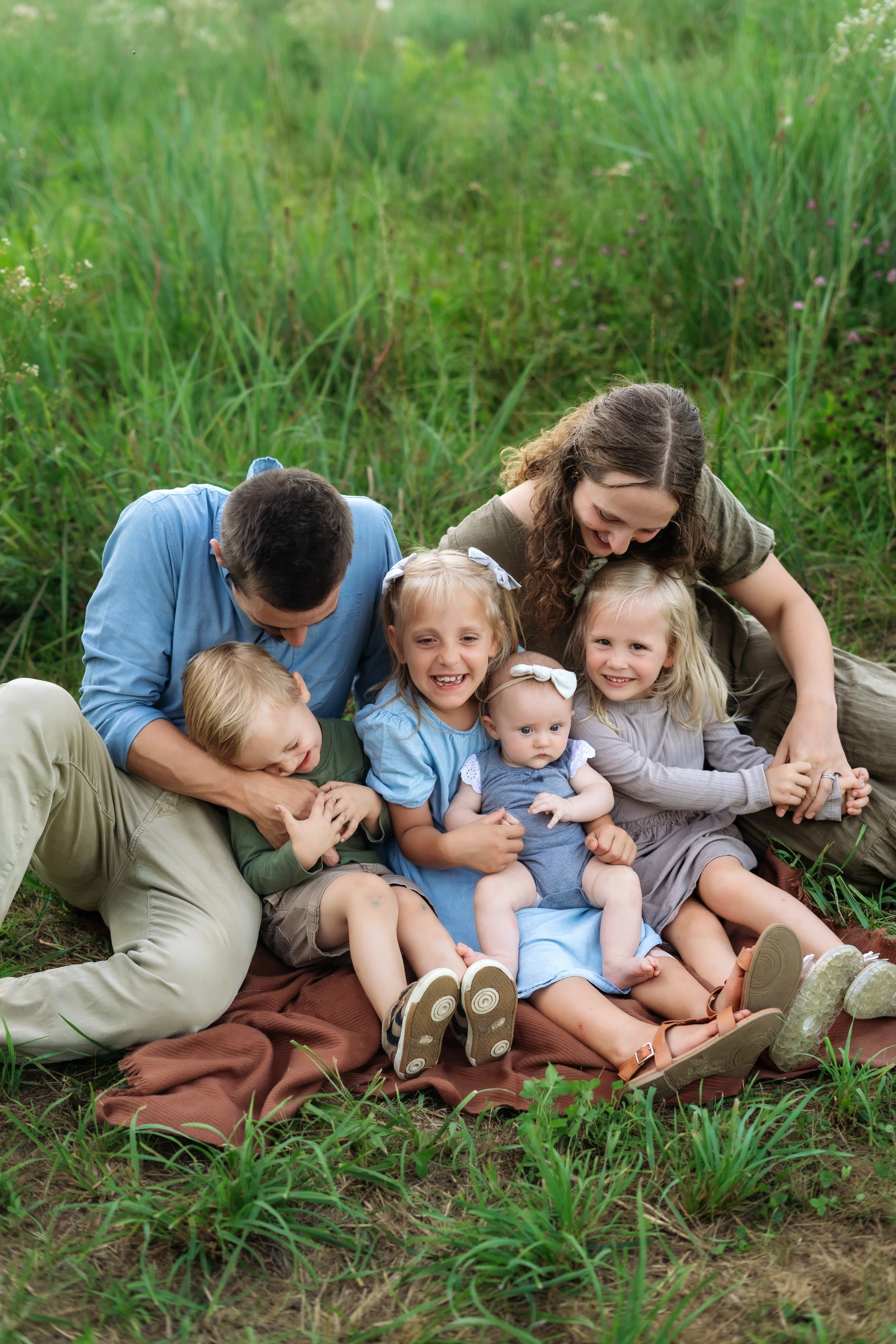 A family with two adults and five children sitting outdoors on a brown blanket in a grassy field, smiling and hugging each other.