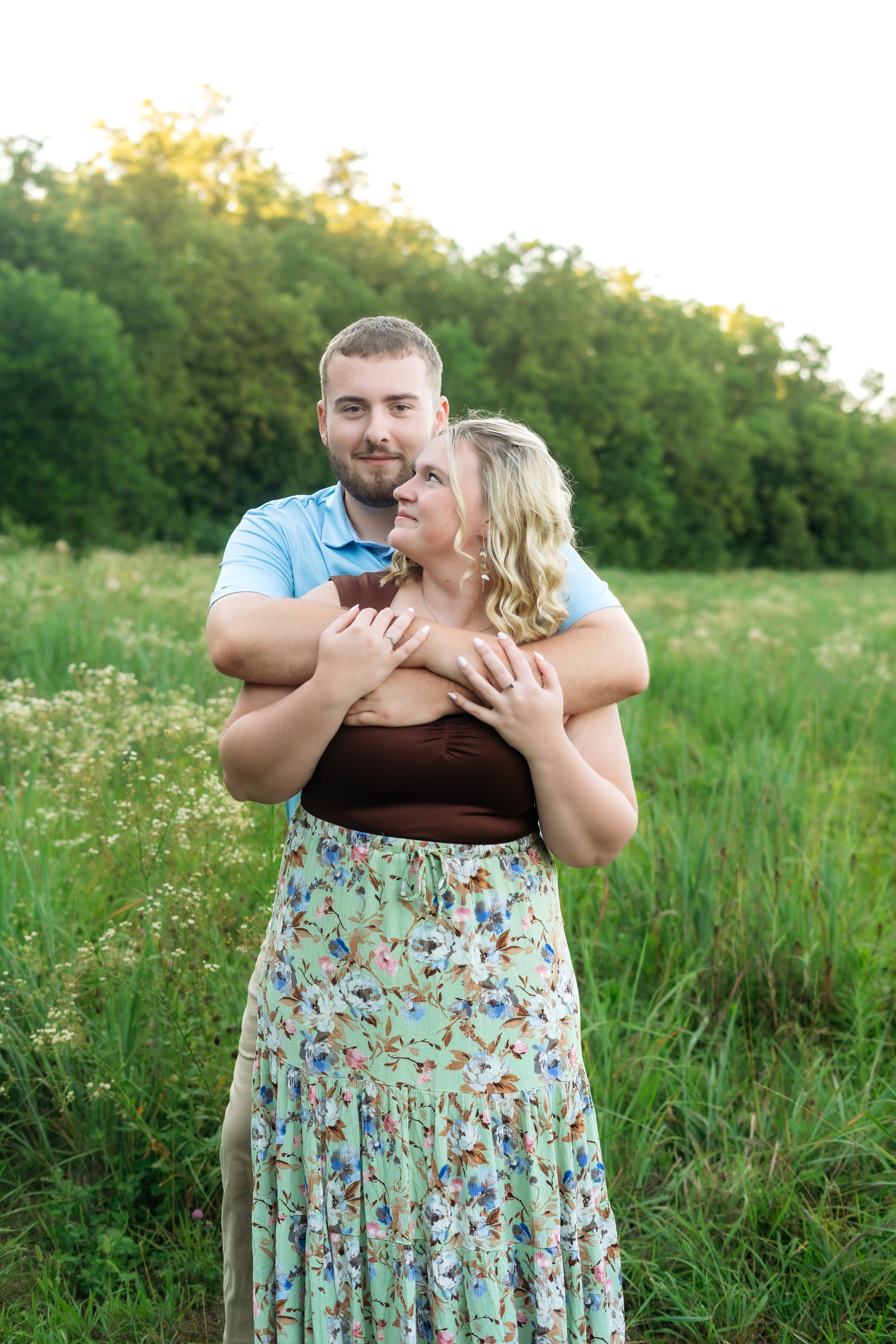 A couple standing in a grassy field with trees in the background. The man is hugging the woman from behind, both smiling and looking content. The woman wears a floral dress, and the man wears a light blue shirt.