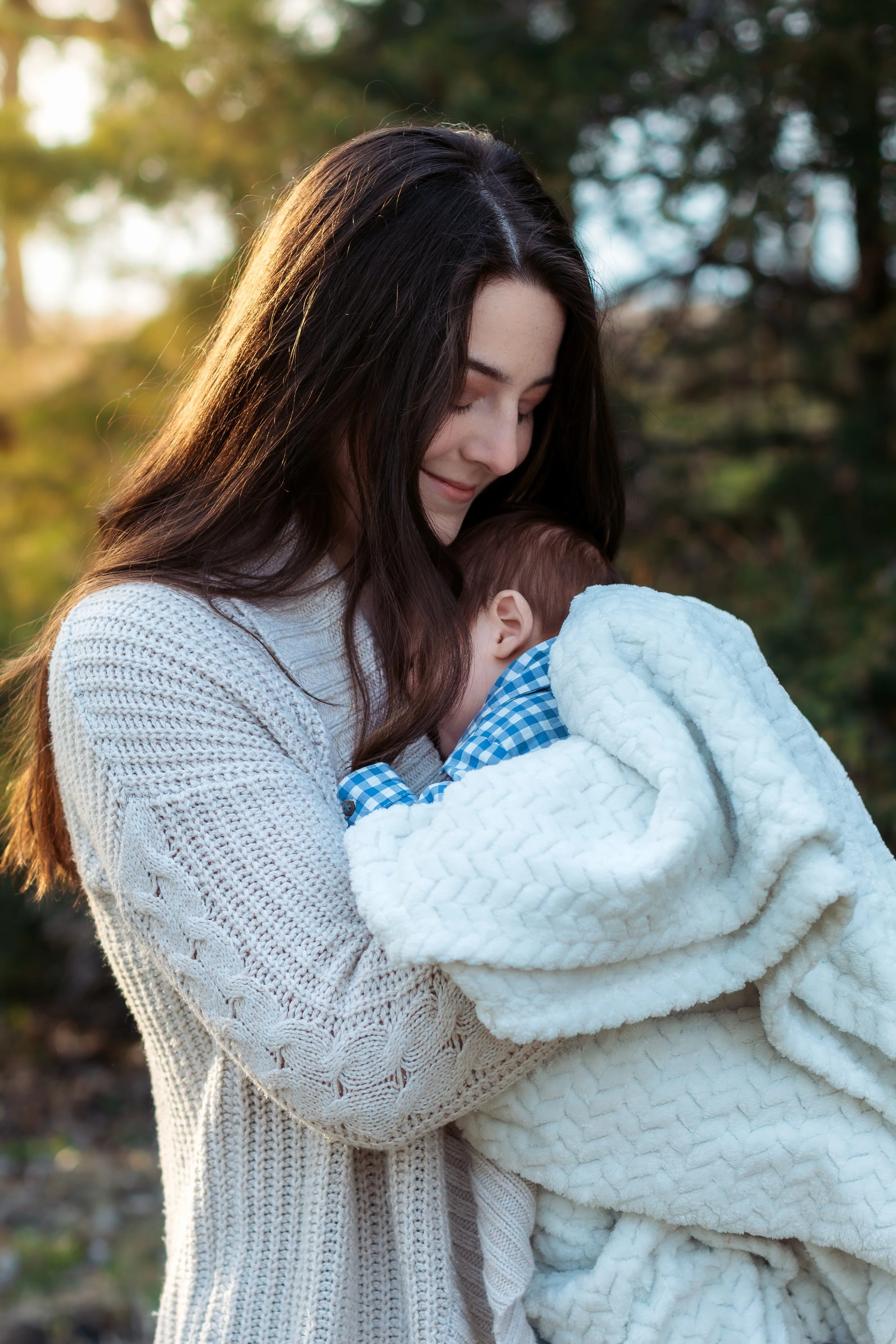 A woman with long dark hair smiling and holding a sleeping baby wrapped in a white blanket outdoors with trees in the background.