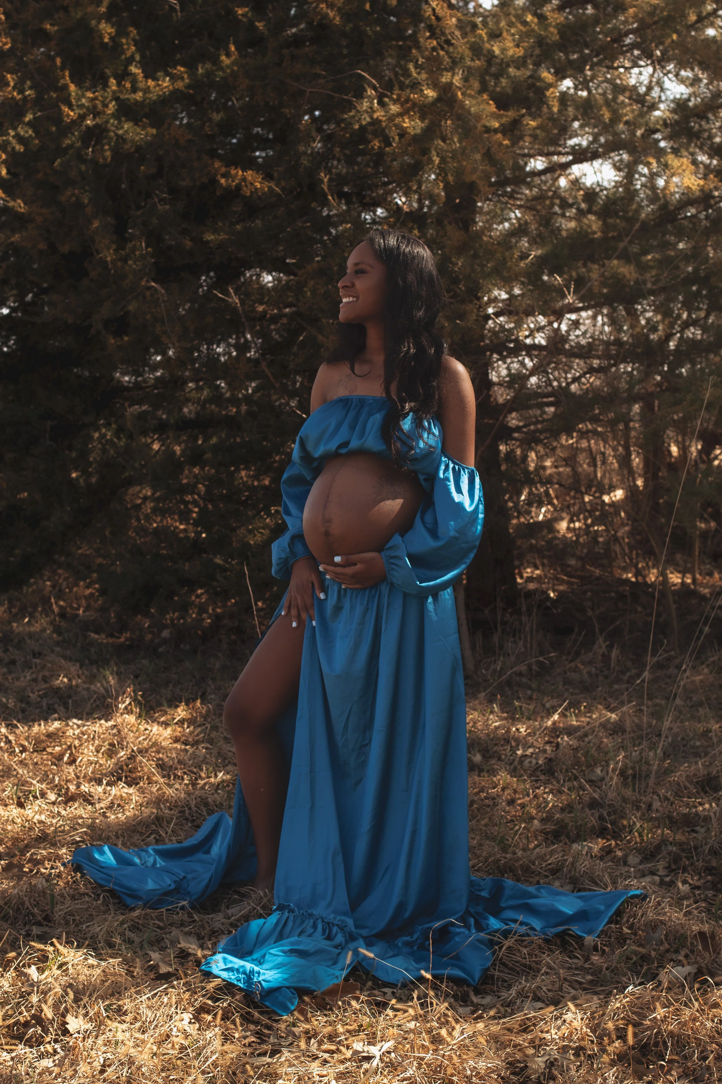 A pregnant woman in a blue dress standing outdoors on dry grass near trees, smiling, with the sunlight creating a warm glow.