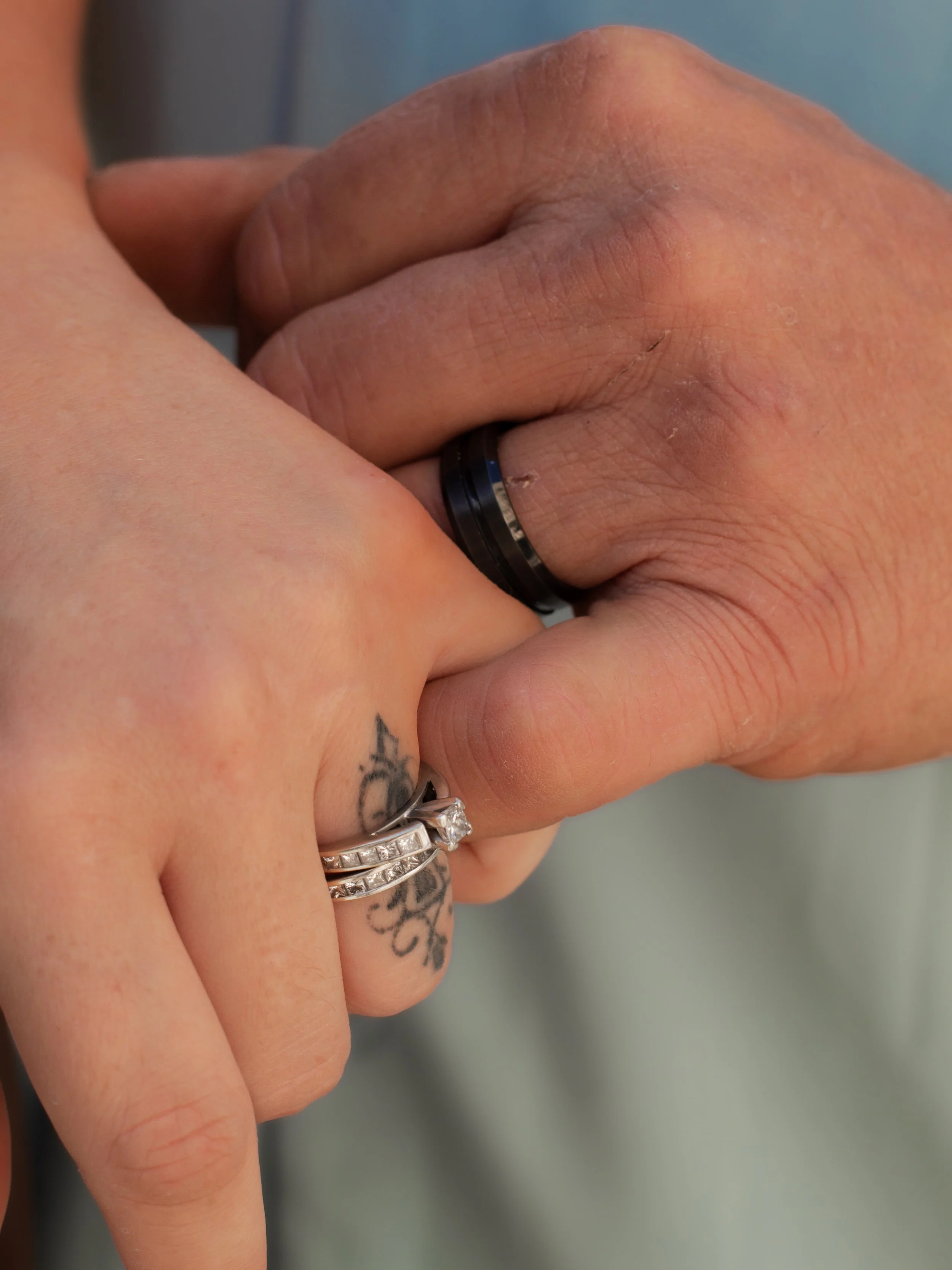 Close-up of two hands, one with a wedding ring and multiple rings, holding each other, displaying detailed skin and a tattoo.