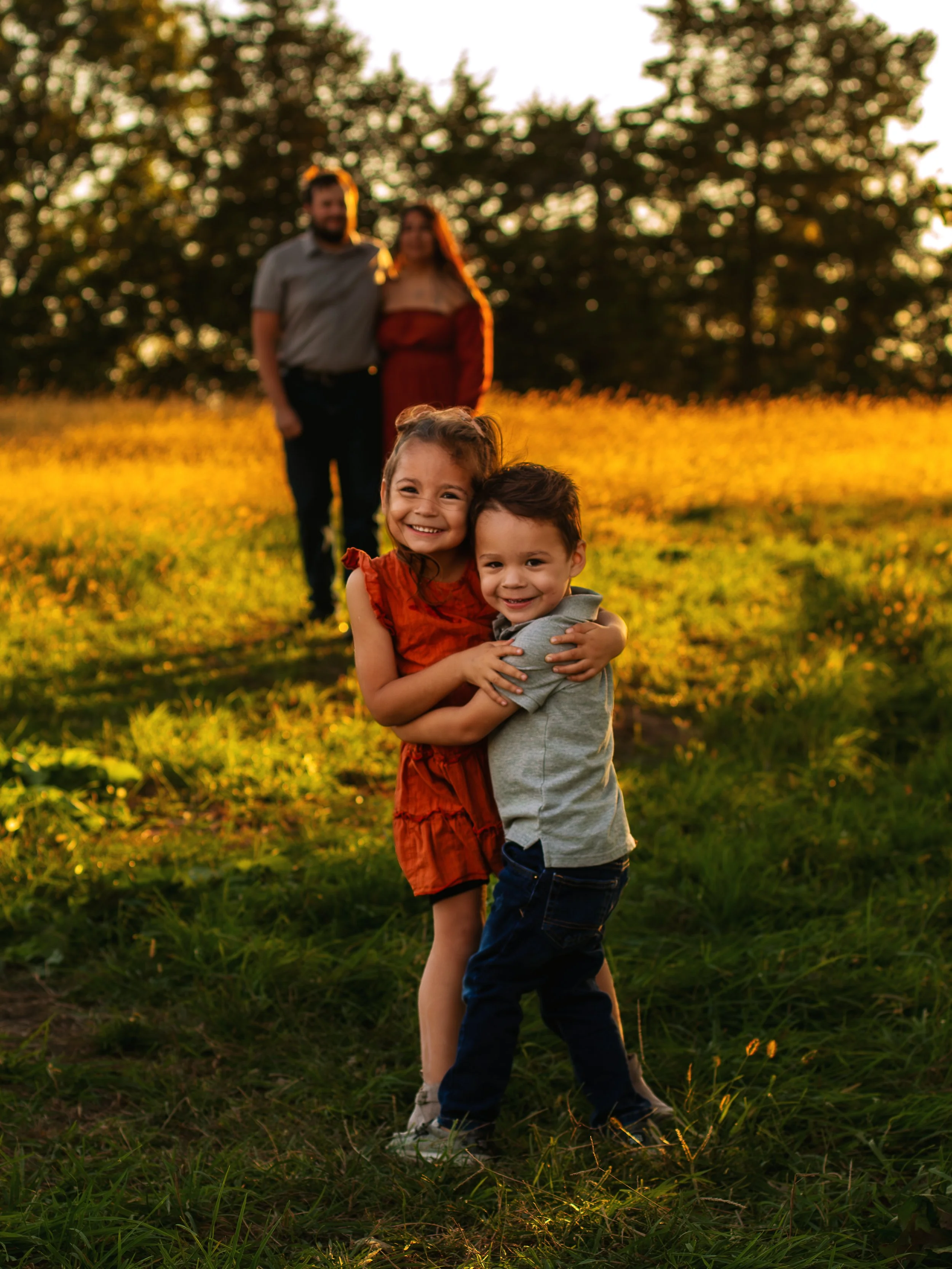 A young girl and boy hugging and smiling at the camera in a field at sunset, with two adults in the background.