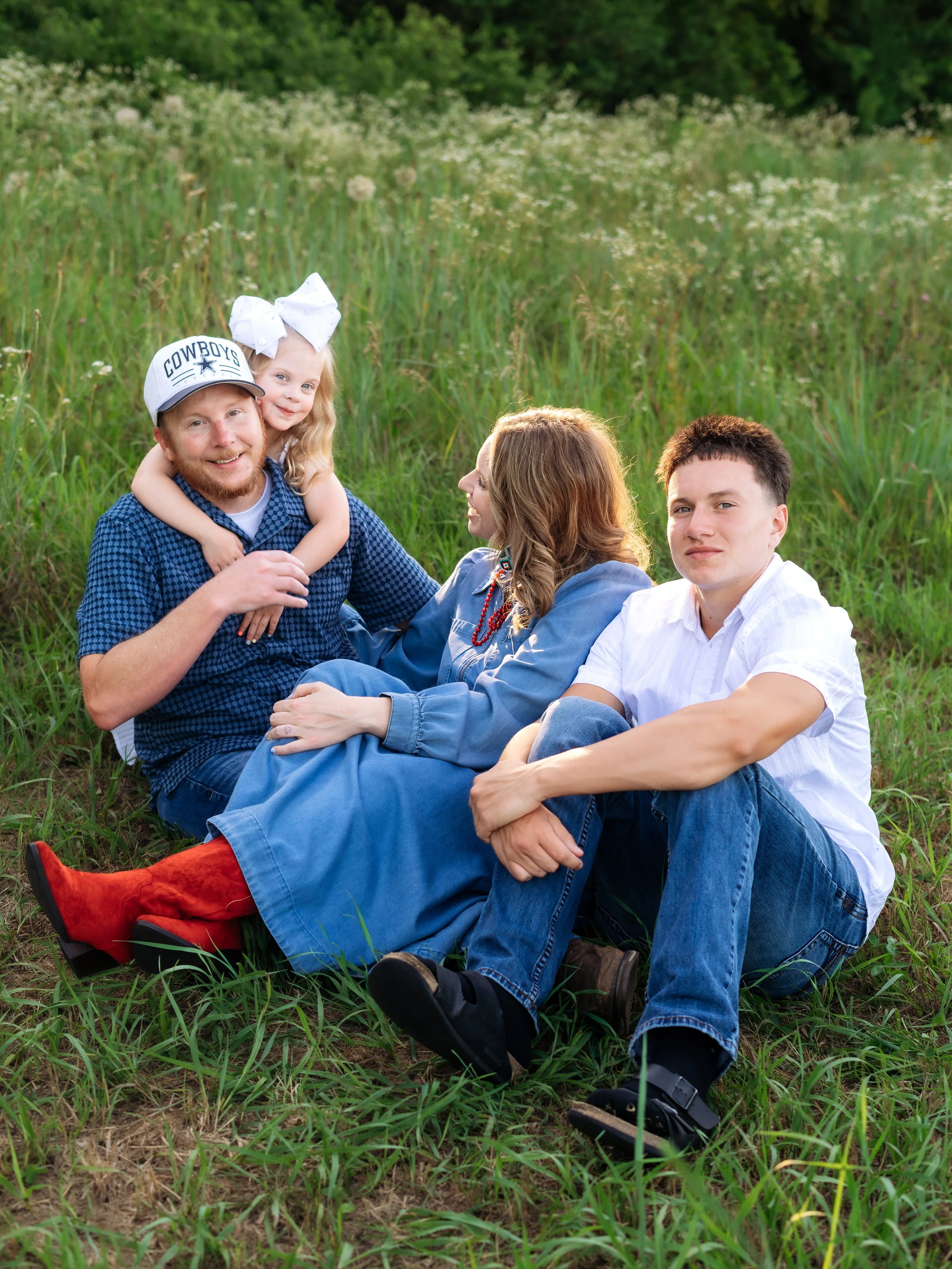 A family of four sitting on the grass in a field, with a man giving a piggyback ride to a young girl wearing a large white bow. The family is outdoors, surrounded by green grass and trees.