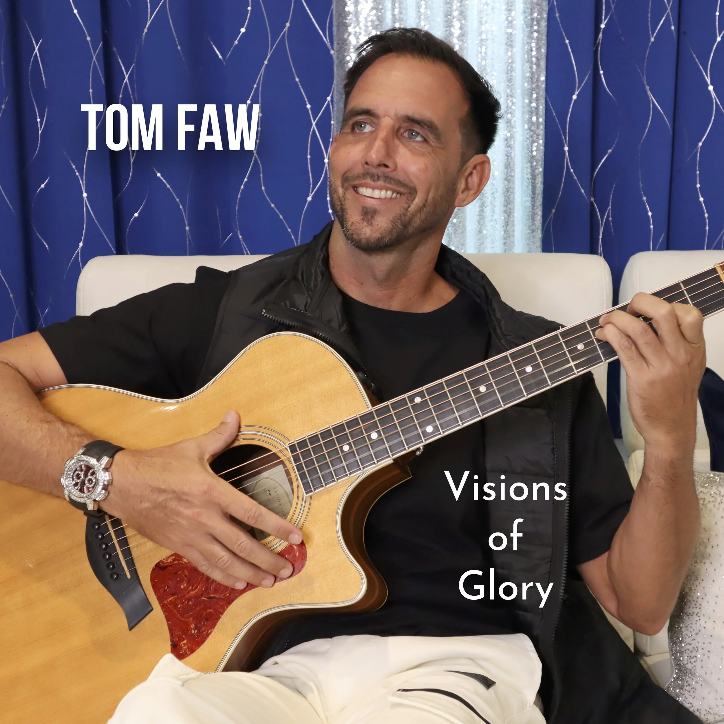 A man with short dark hair and a goatee playing an acoustic guitar, smiling, sitting on a white chair in front of a blue curtain with silver and white star-like decorations, wearing a black shirt and a watch with a large face.